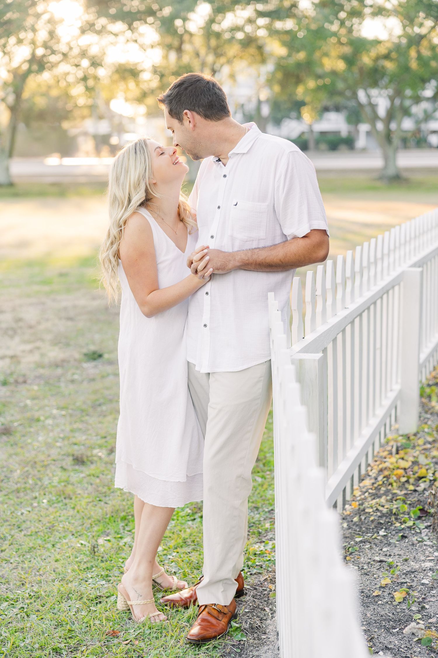 A couple dressed in white shares playful moments near a picket fence during sunset.
