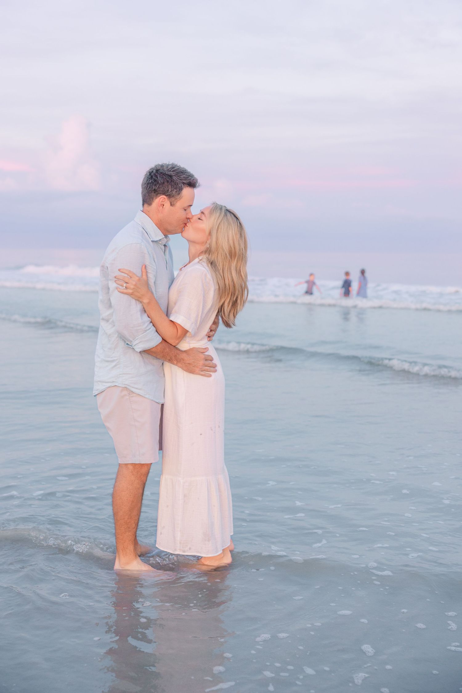 A romantic embrace in the shallow ocean water during sunset with soft pink skies.