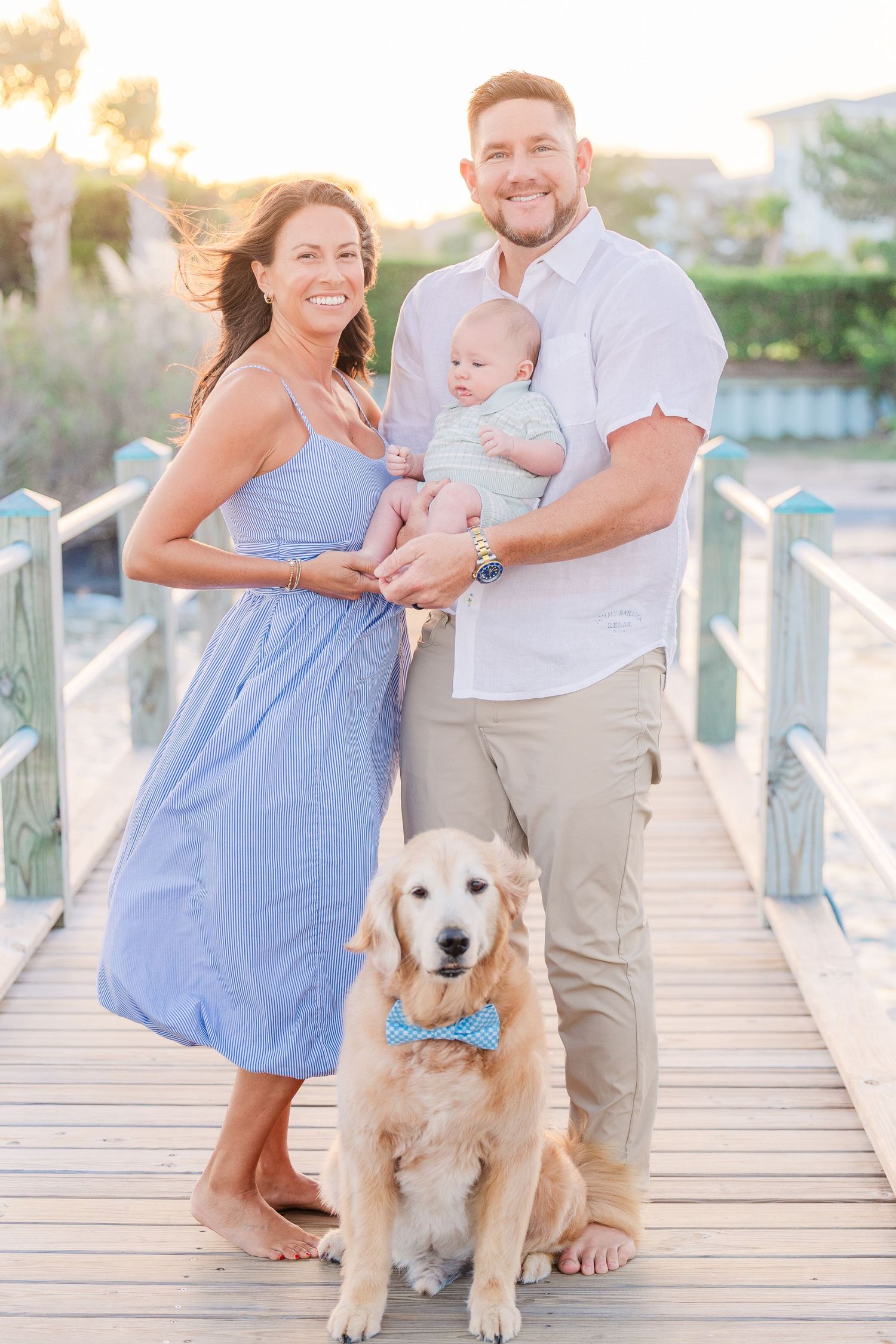 A happy family poses with their golden retriever wearing a blue bowtie on a coastal boardwalk.