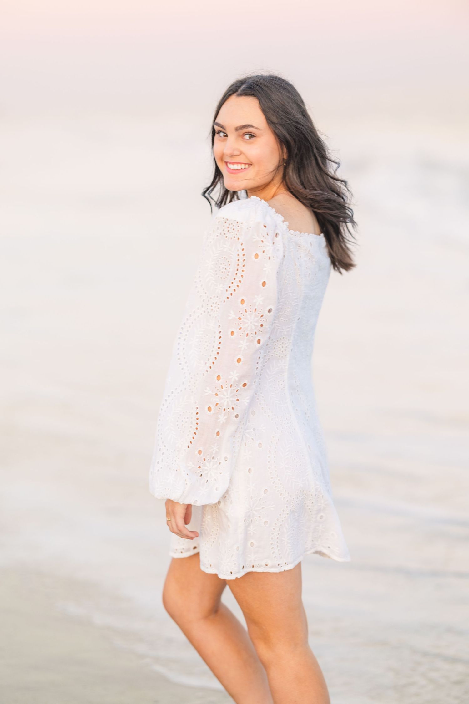 White eyelet dress with long sleeves photographed against pastel sunset sky at the beach.