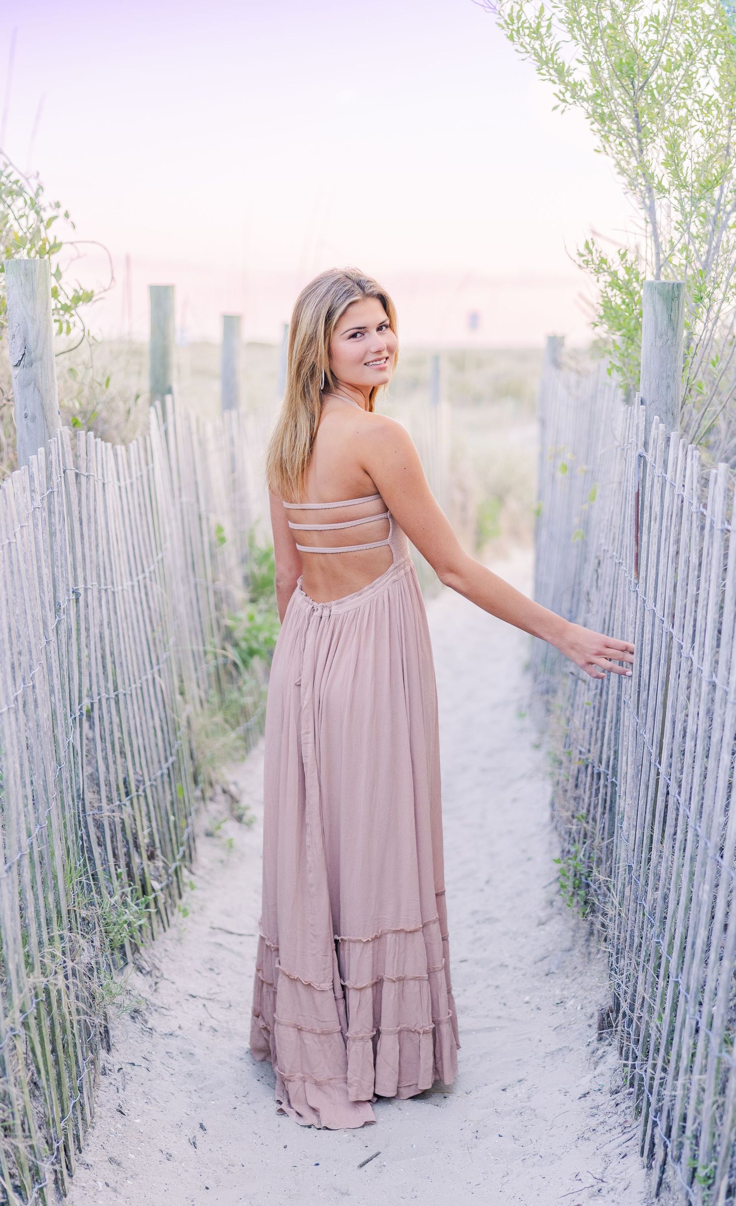 Woman in a flowing dusty rose dress poses gracefully along a rustic beach fence at golden hour.