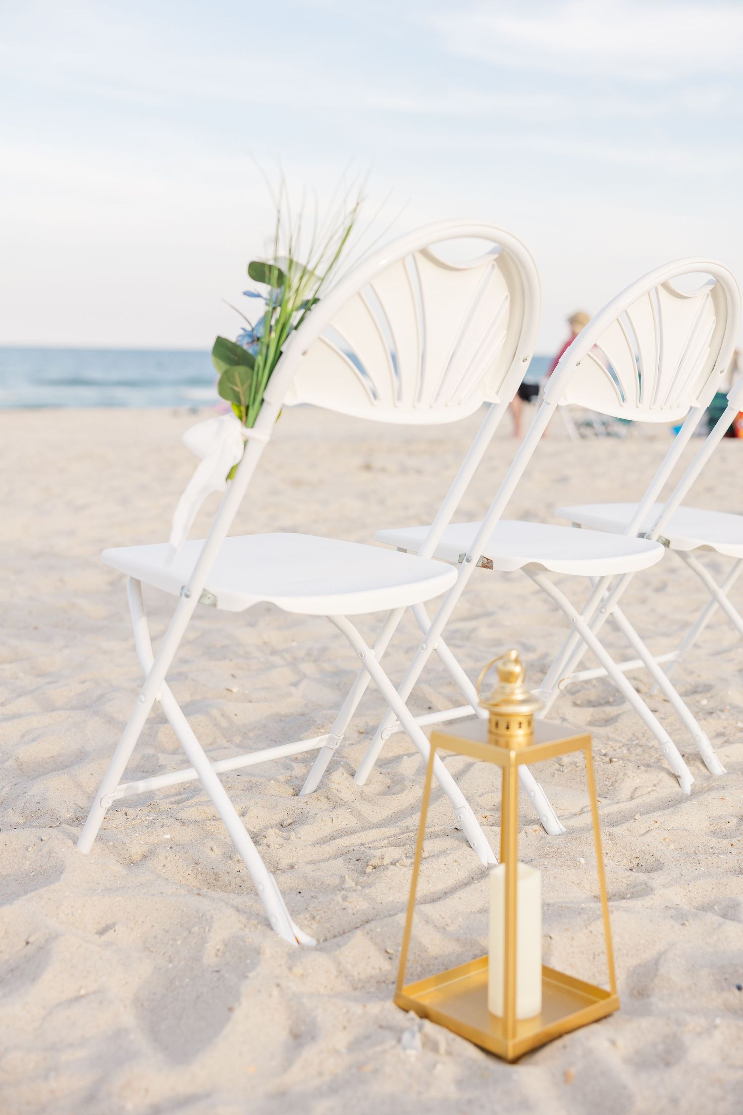 White folding chairs with greenery and gold lantern on sandy beach wedding ceremony setup in Carolina Beach near Wilmington, NC.