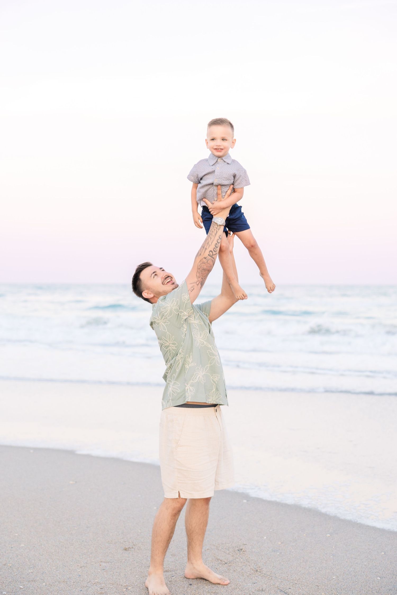 Father lifting young son high above his head on a sandy beach at sunset with ocean waves in the background.