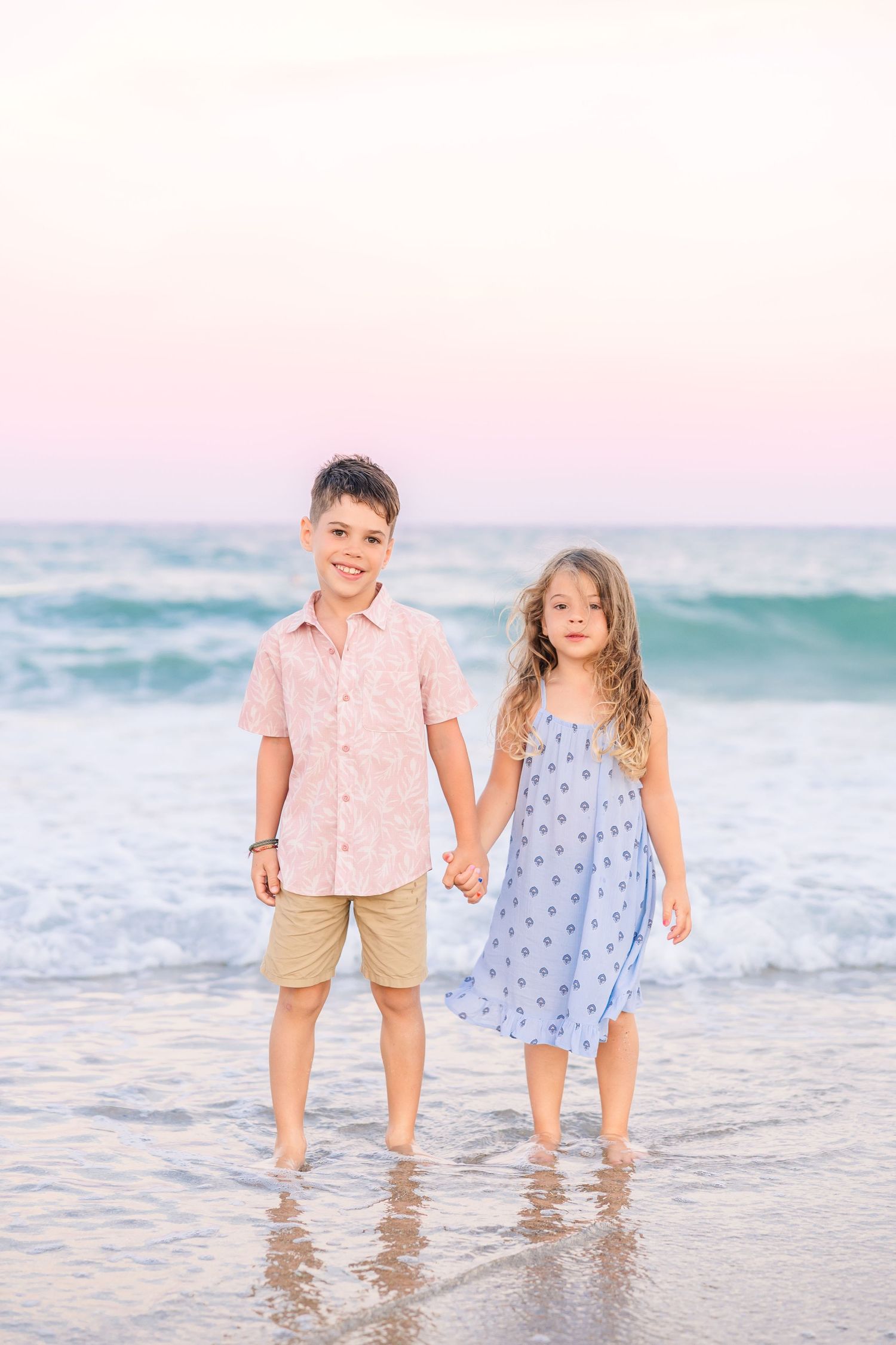 A young boy and girl hold hands while standing in the ocean waves on a beach at sunset.