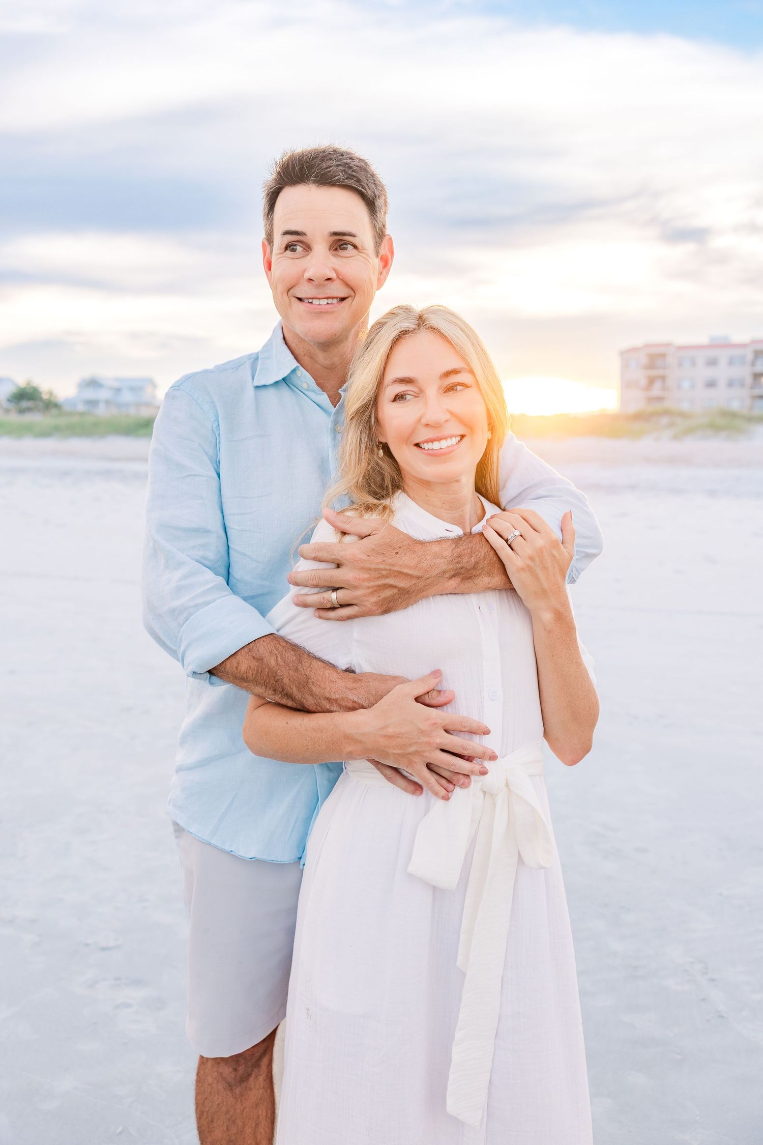 Couple sharing intimate moments on sandy beach at sunset with coastal buildings visible.