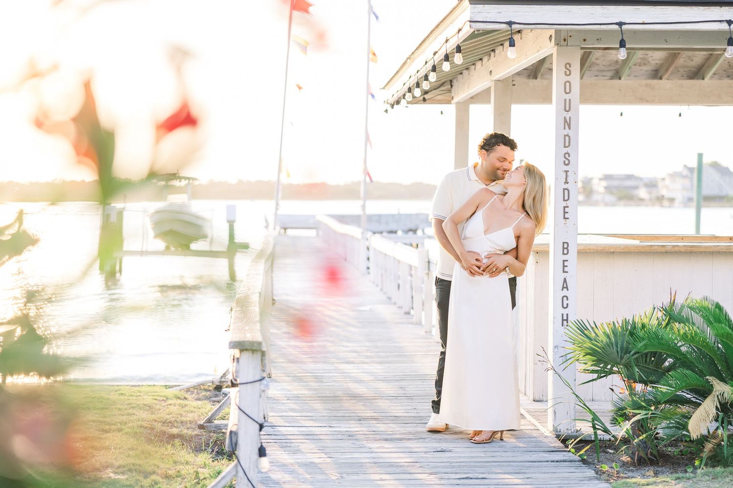 Couple sharing a romantic embrace on a waterfront dock at golden hour with soft bokeh flowers in foreground.