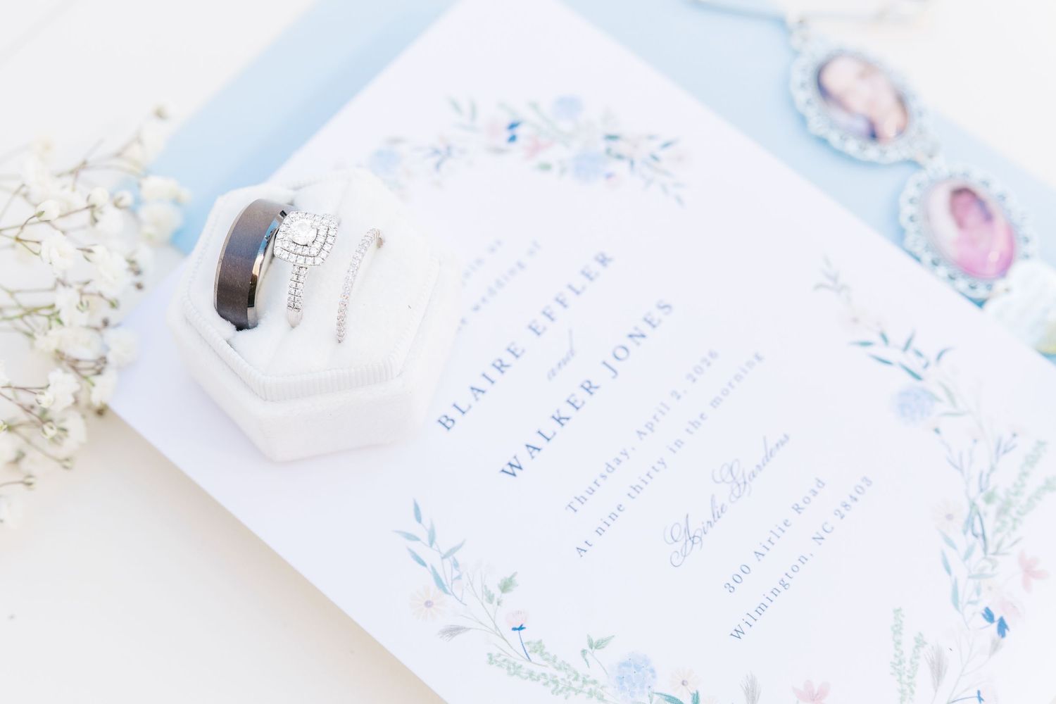 Elegant wedding invitation with blue floral border, diamond engagement ring, and wedding band in a white velvet ring box in Airlie Gardens i