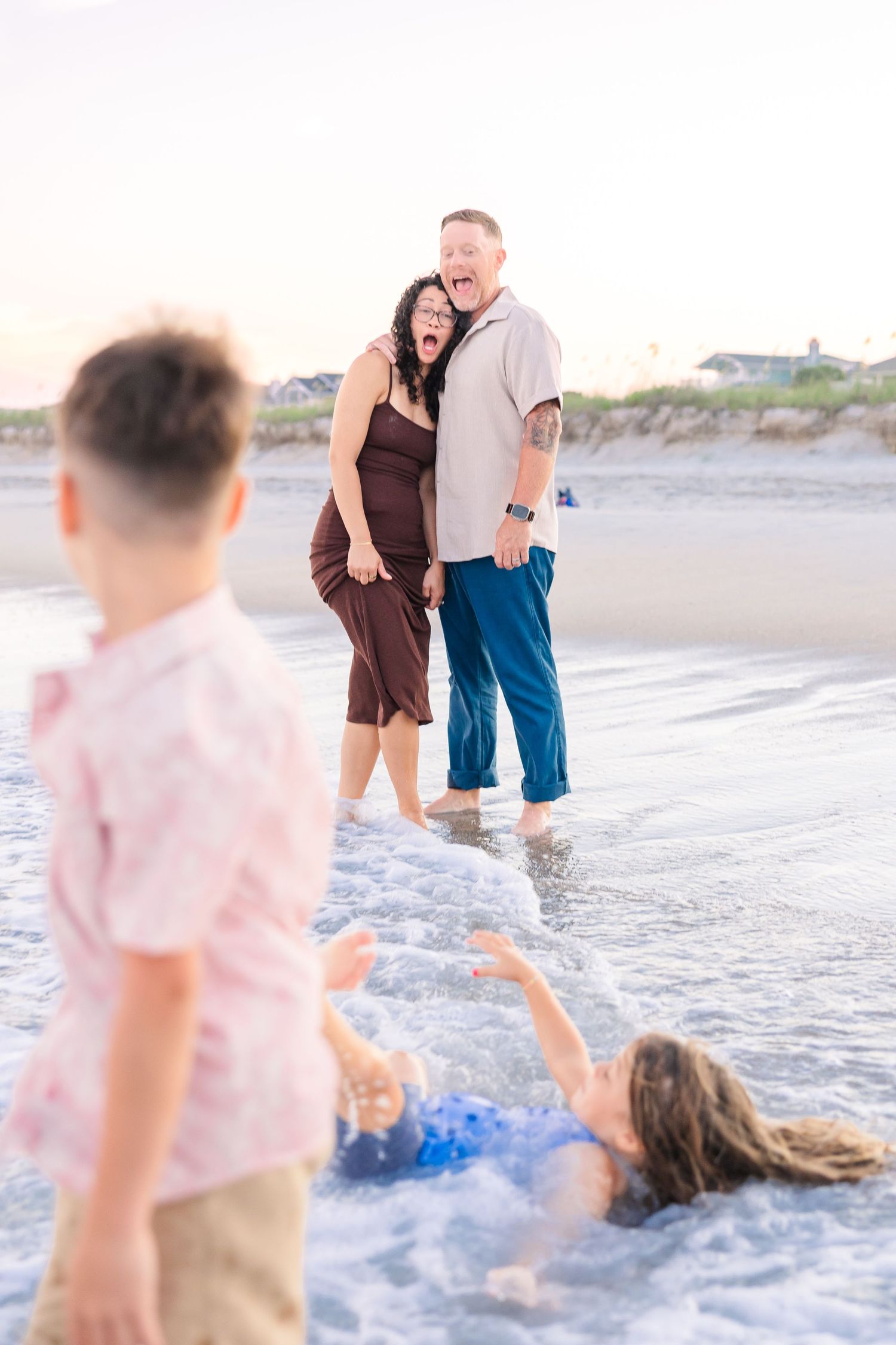 A couple laughs as their children play in the ocean waves during a family beach photoshoot.