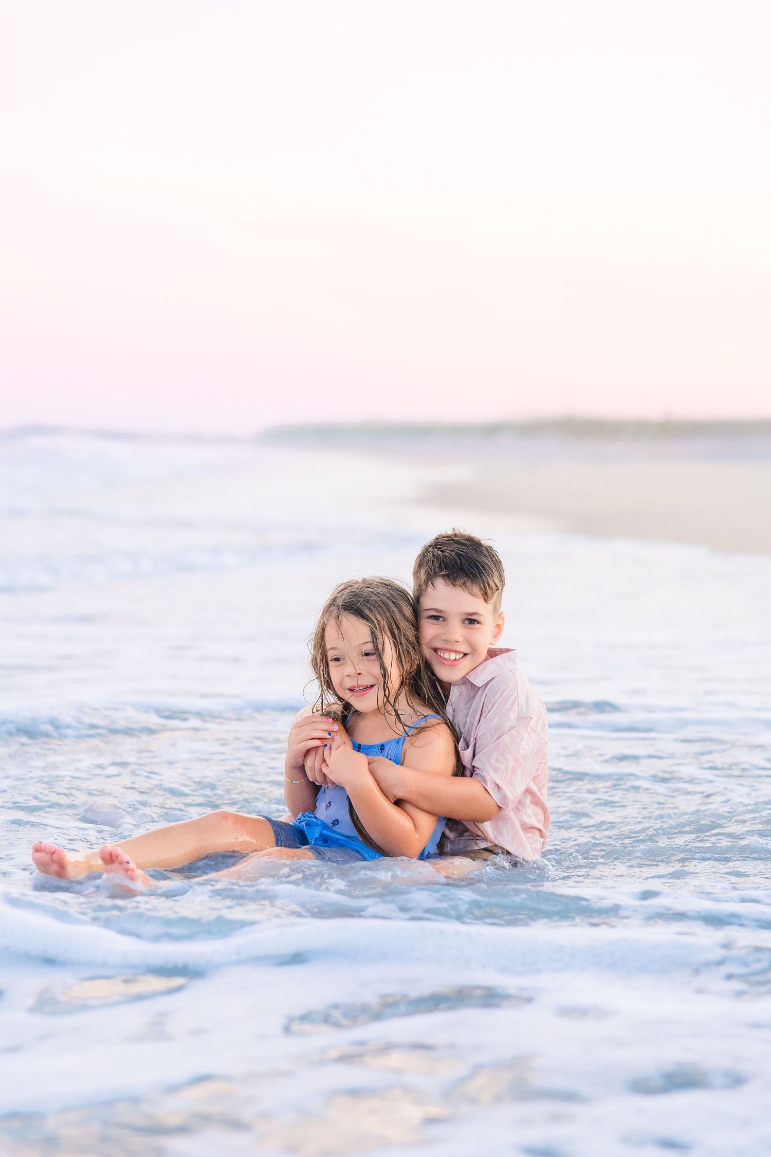 Two happy siblings hug while sitting in ocean waves on a beach at sunset.