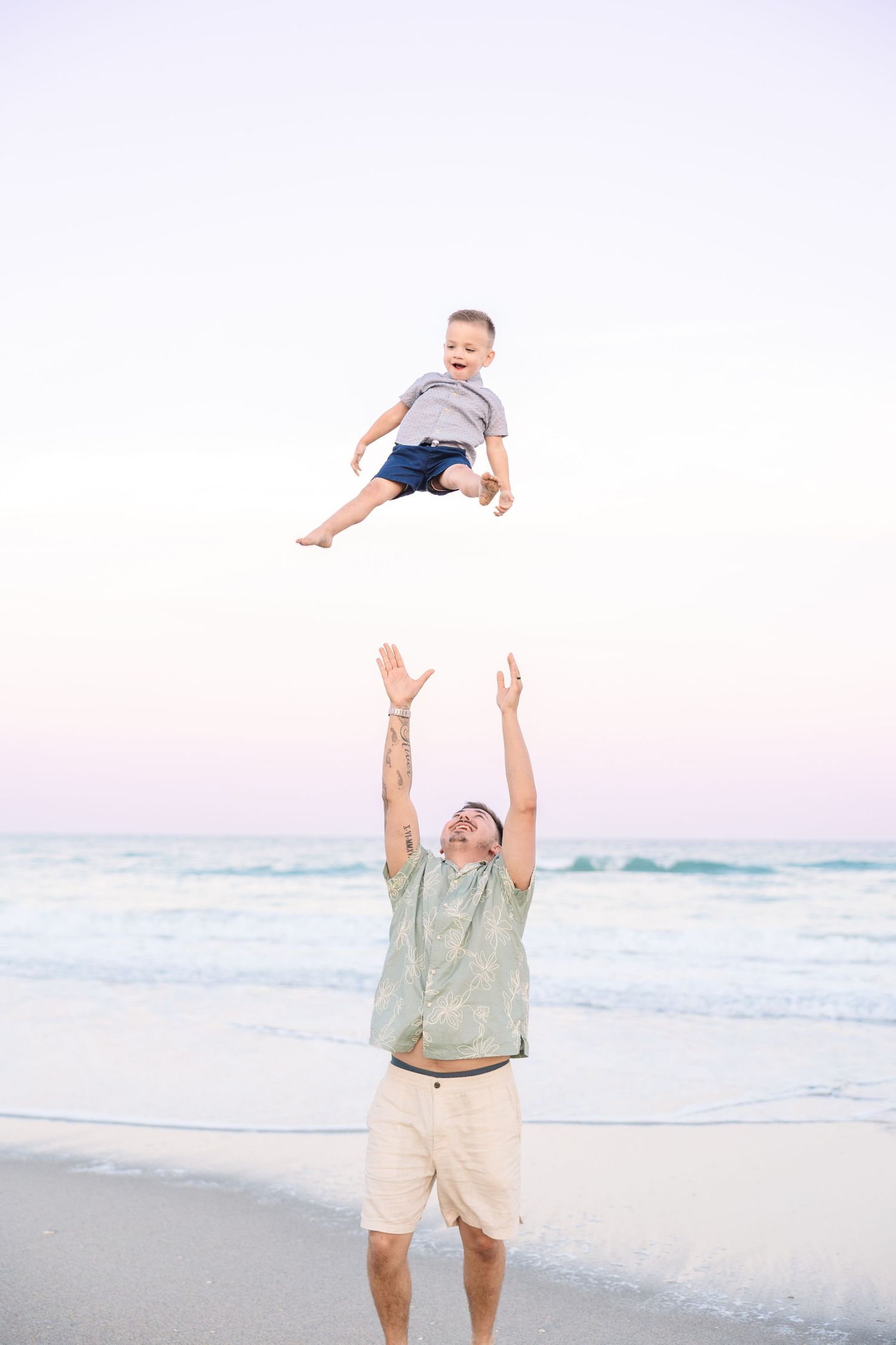 Father tossing smiling toddler son into the air on a beach at dusk with ocean waves behind them.