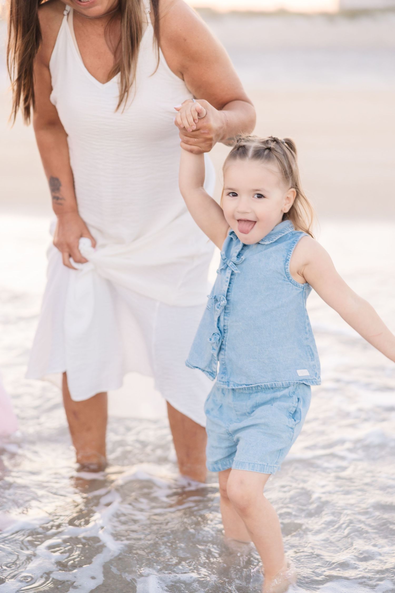 Adorable toddler girl in denim romper laughing while holding moms hand in beach waves.