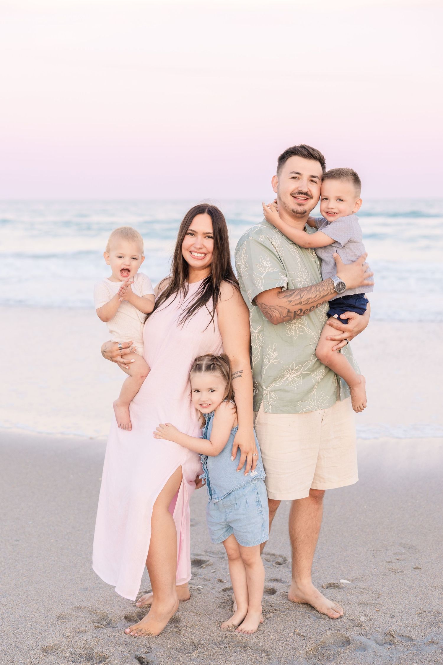 Happy family of five posing together on a sandy beach at sunset, parents holding toddlers with child standing between them.