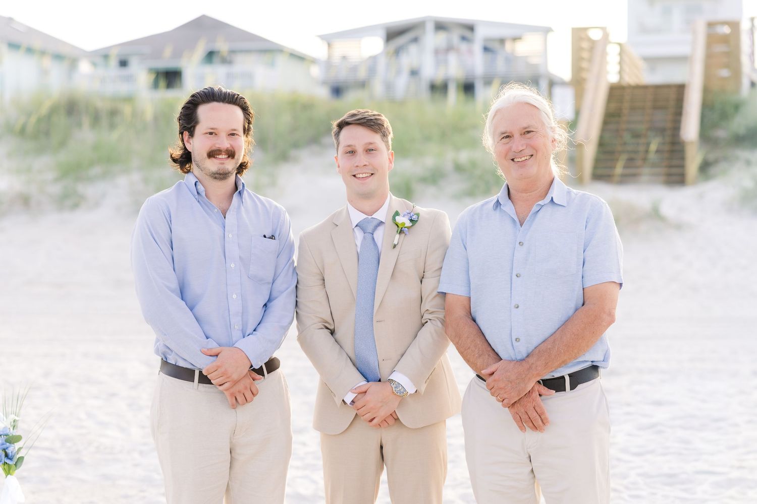 Three men smile on a sandy beach at a wedding, with the groom in a tan suit flanked by two guests in blue shirts in Carolina Beach near Wilm
