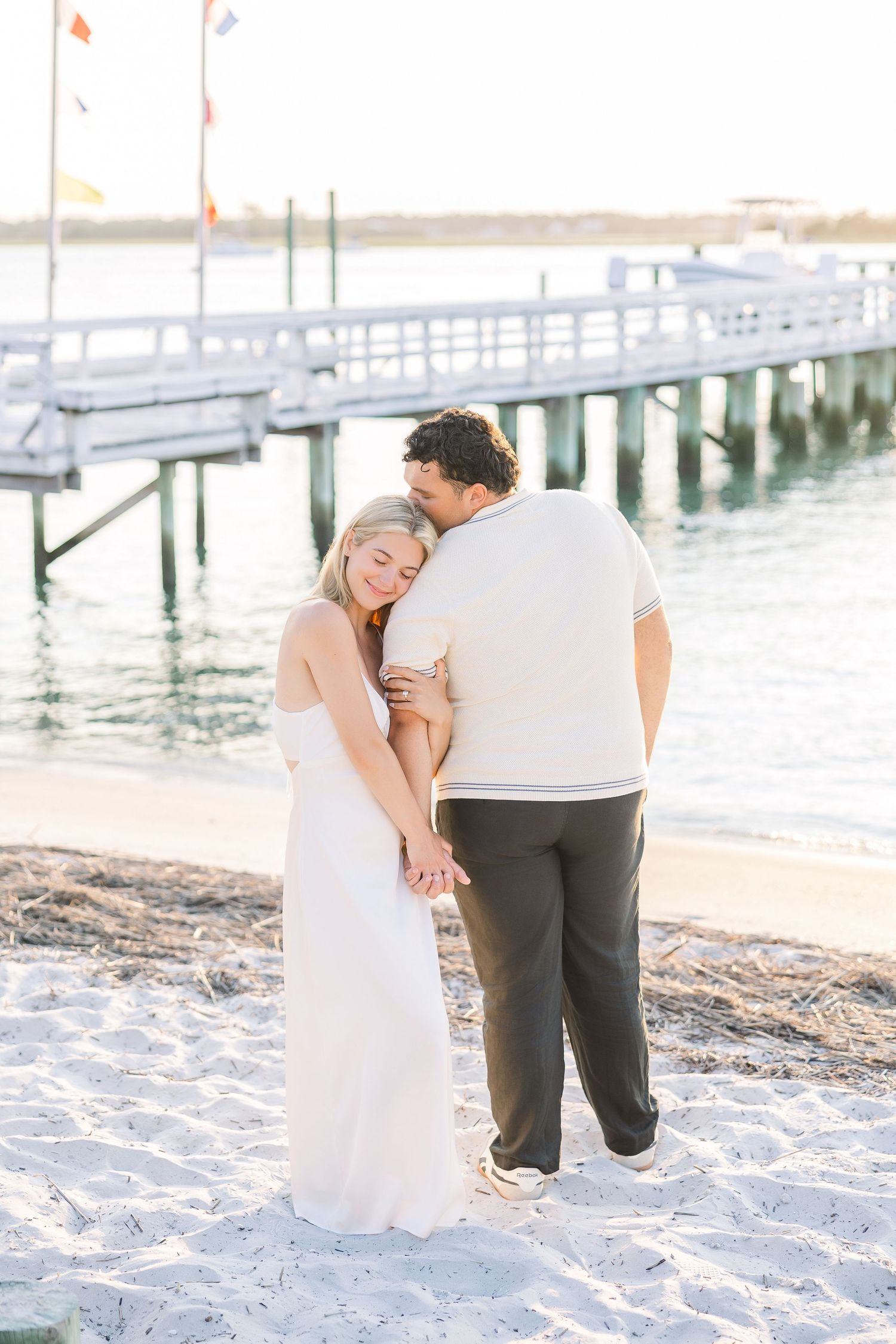 Romantic couple embracing on sandy beach near pier at sunset, woman in white dress.