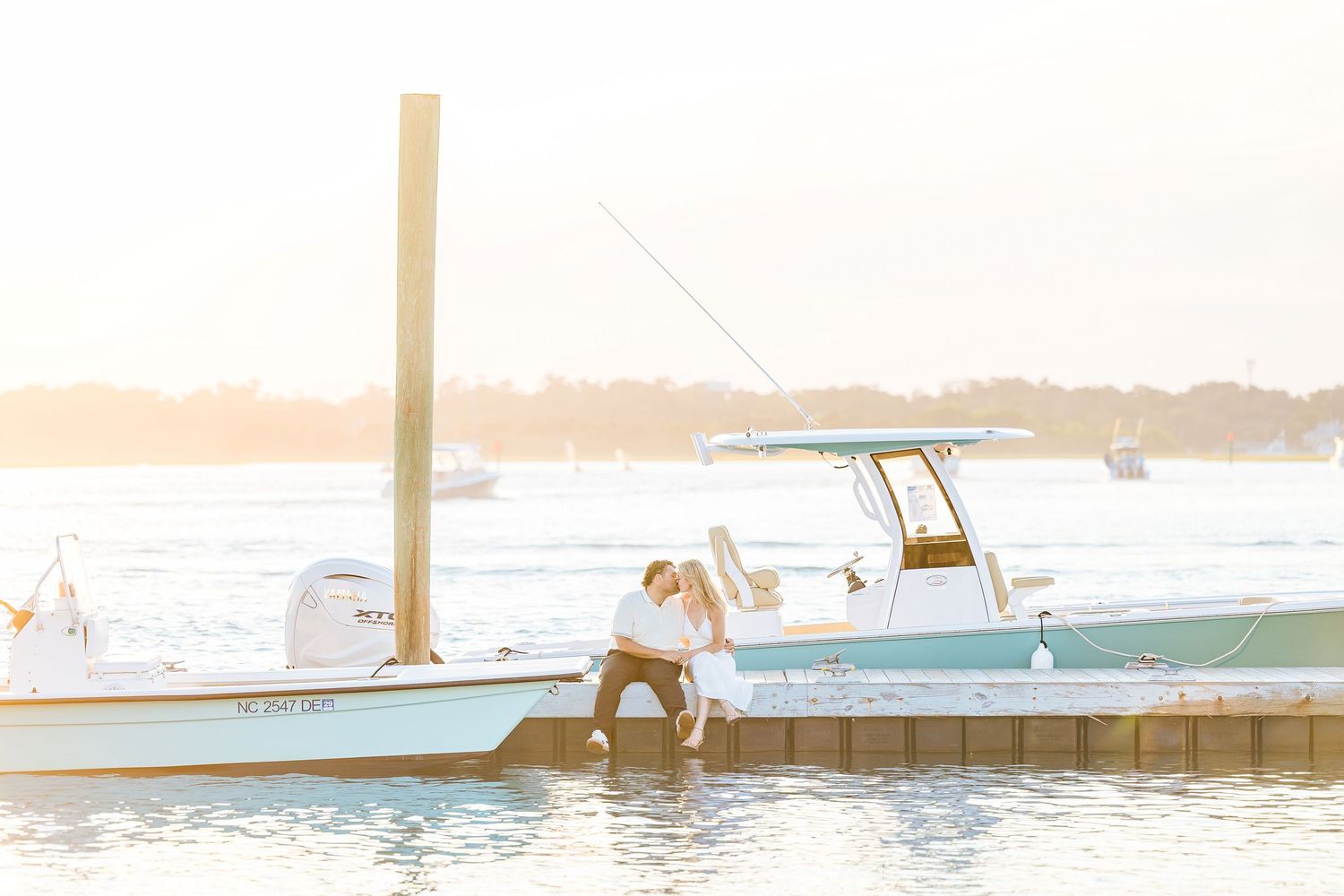 Couple sharing a romantic kiss on a wooden dock beside a white boat at golden hour sunset on calm waters.