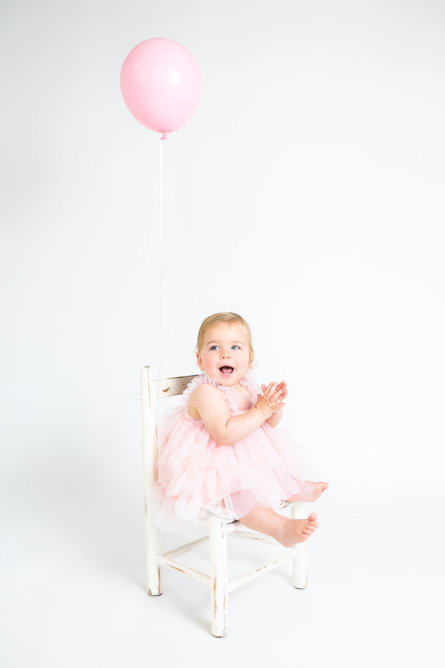 Baby in pink dress playing with balloon while sitting on white chair.