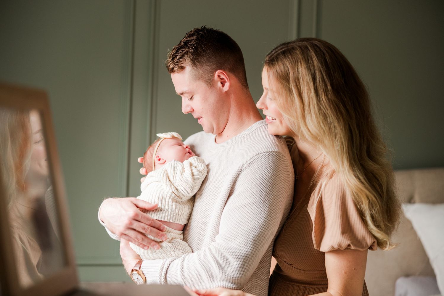 Parents holding newborn baby in naturally lit room with green walls.