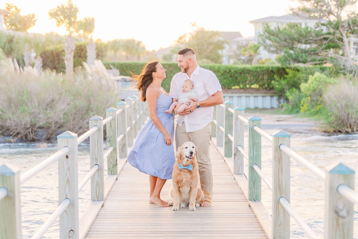 Family and golden retriever dog pose together on wooden boardwalk at sunset near water.