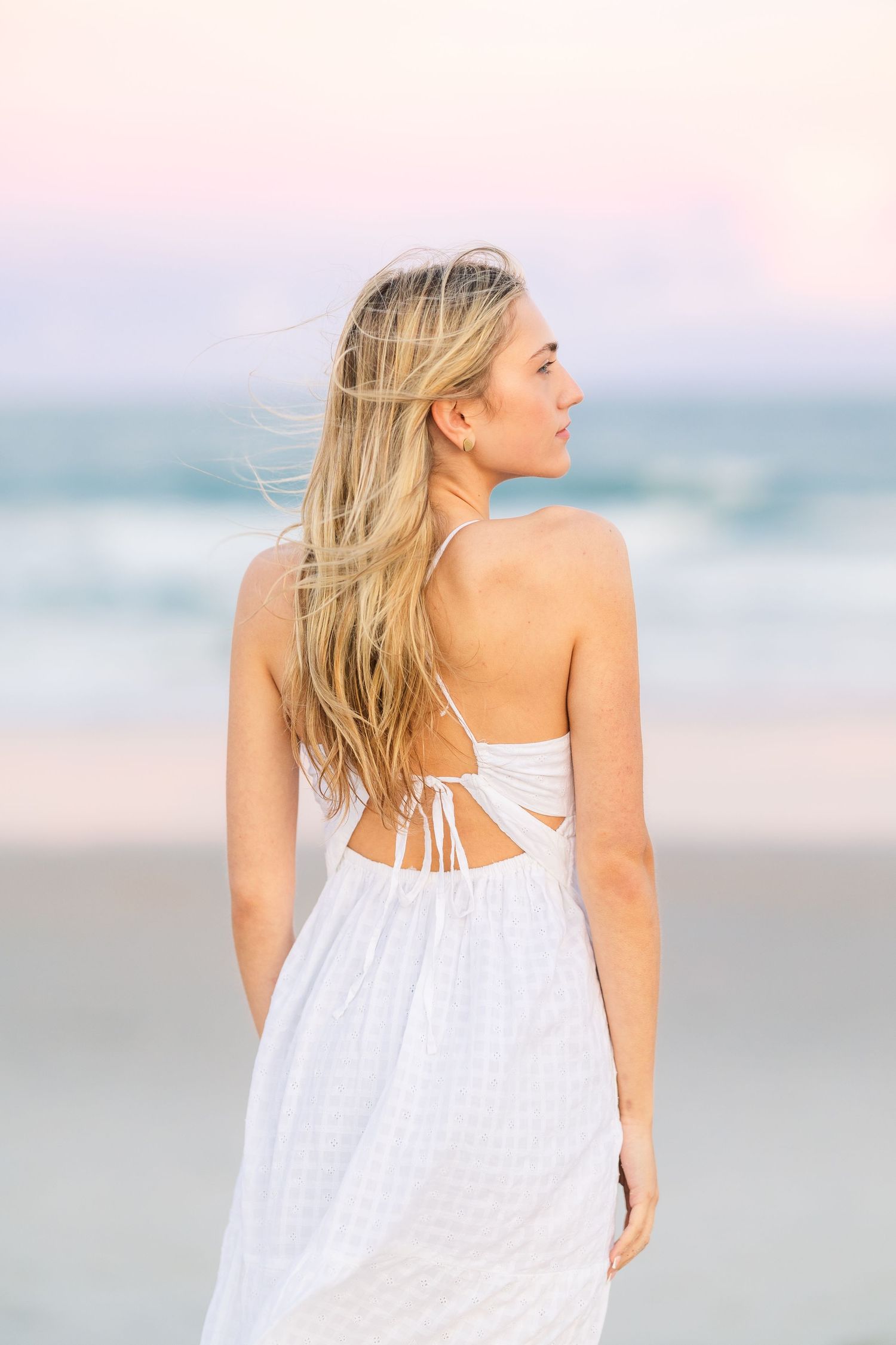 A person in a white summer dress shows the open-back detail while standing on a beach at dusk.