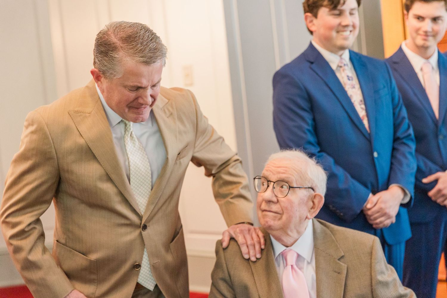 Men in suits share a touching moment with an elderly gentleman at a formal event.