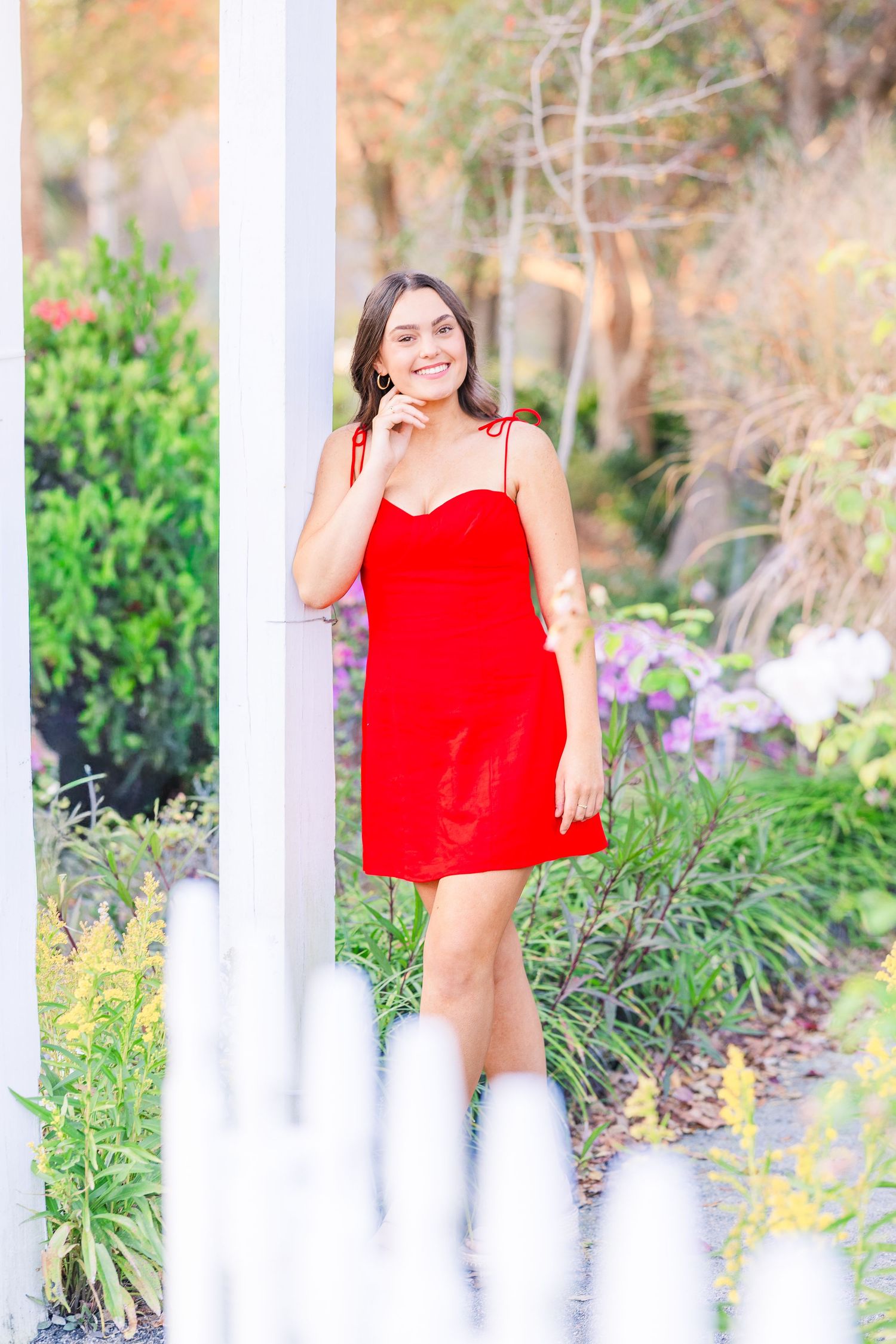Outdoor portrait by white column with red dress and garden flowers in background.