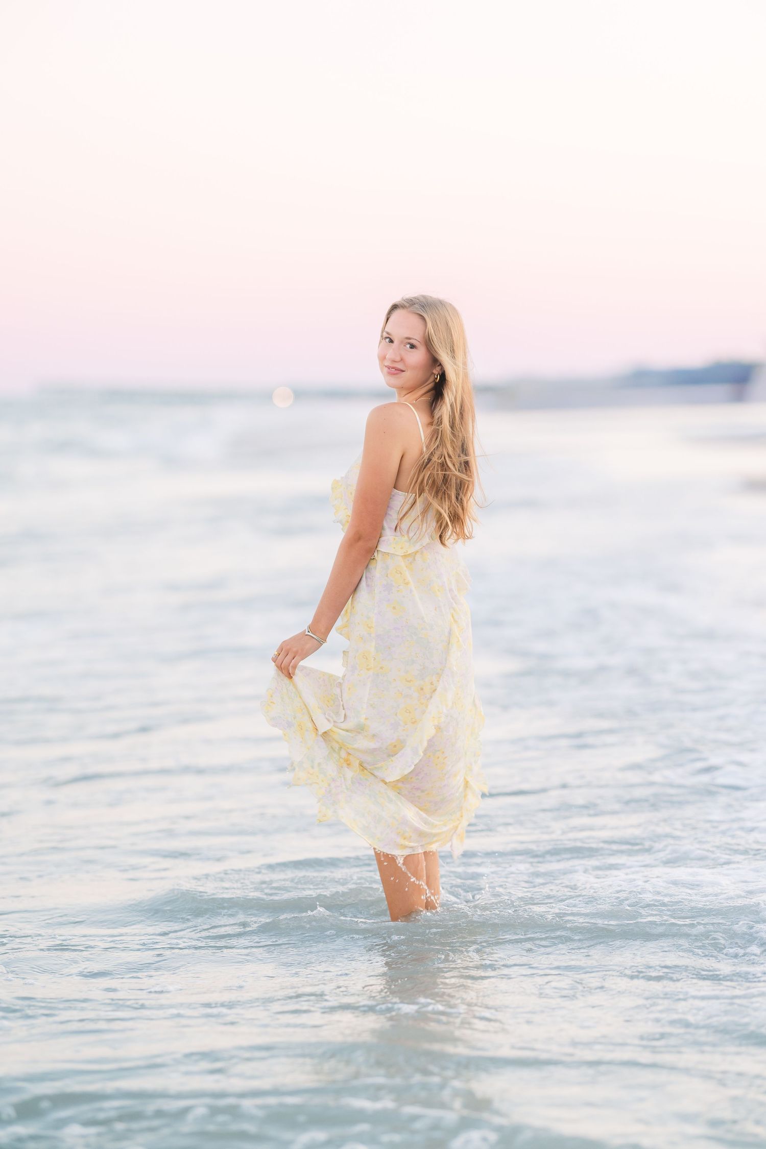 Figure in a light dress wades through calm ocean waters during golden hour.