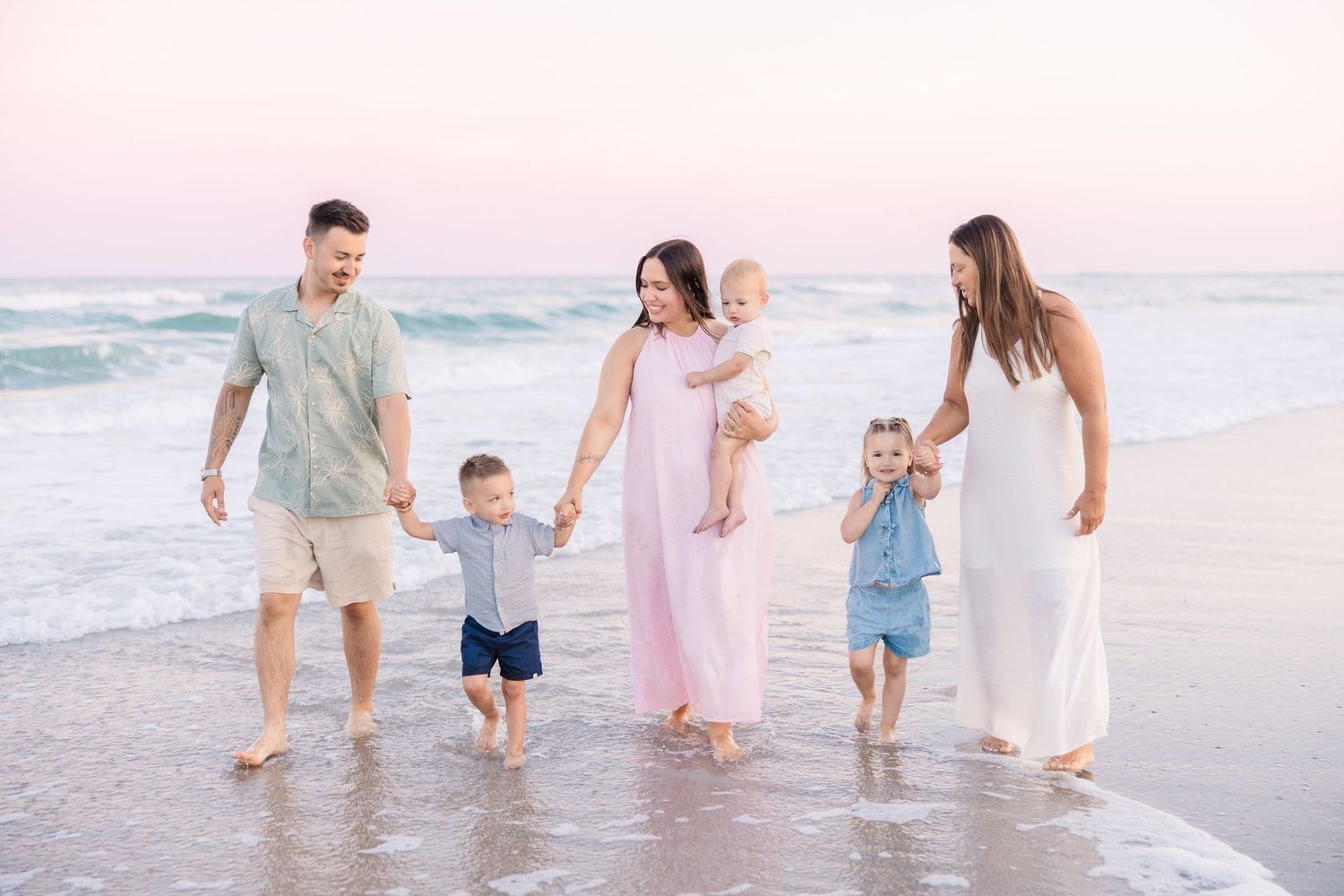 Family of five holding hands walking along beach shoreline at sunset with pink sky backdrop.