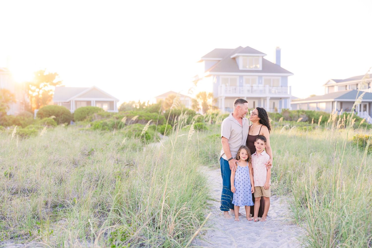 Family of five posing on a sandy beach path at golden hour with coastal homes and tall grass in the background.