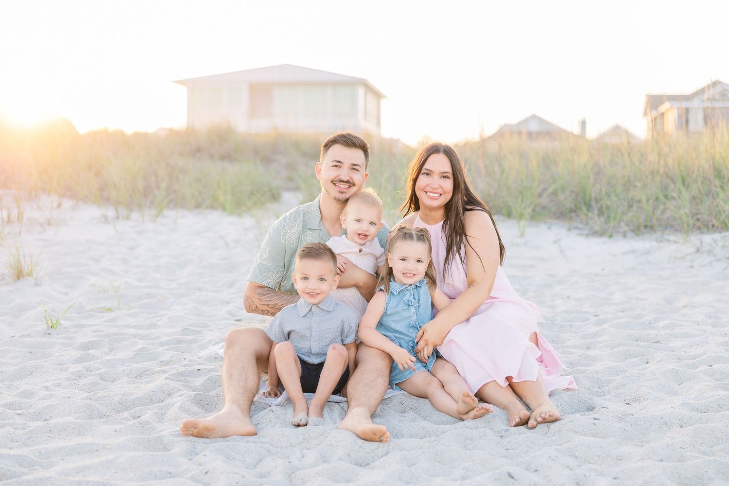 Happy family of five sitting together on sandy beach at sunset, smiling for portrait.
