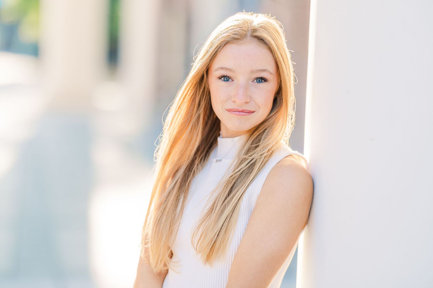 Young woman with long blonde hair poses outdoors in natural sunlight wearing a white sleeveless top.