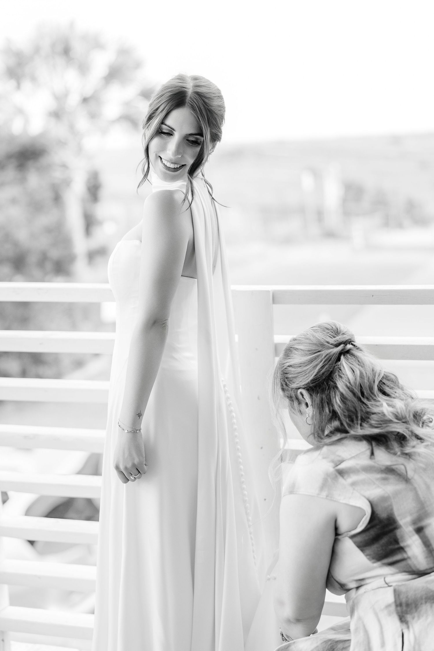 Black and white photo of a bride smiling as bridesmaid fastens her white halter wedding gown on a balcony in Carolina Beach near Wilmington,