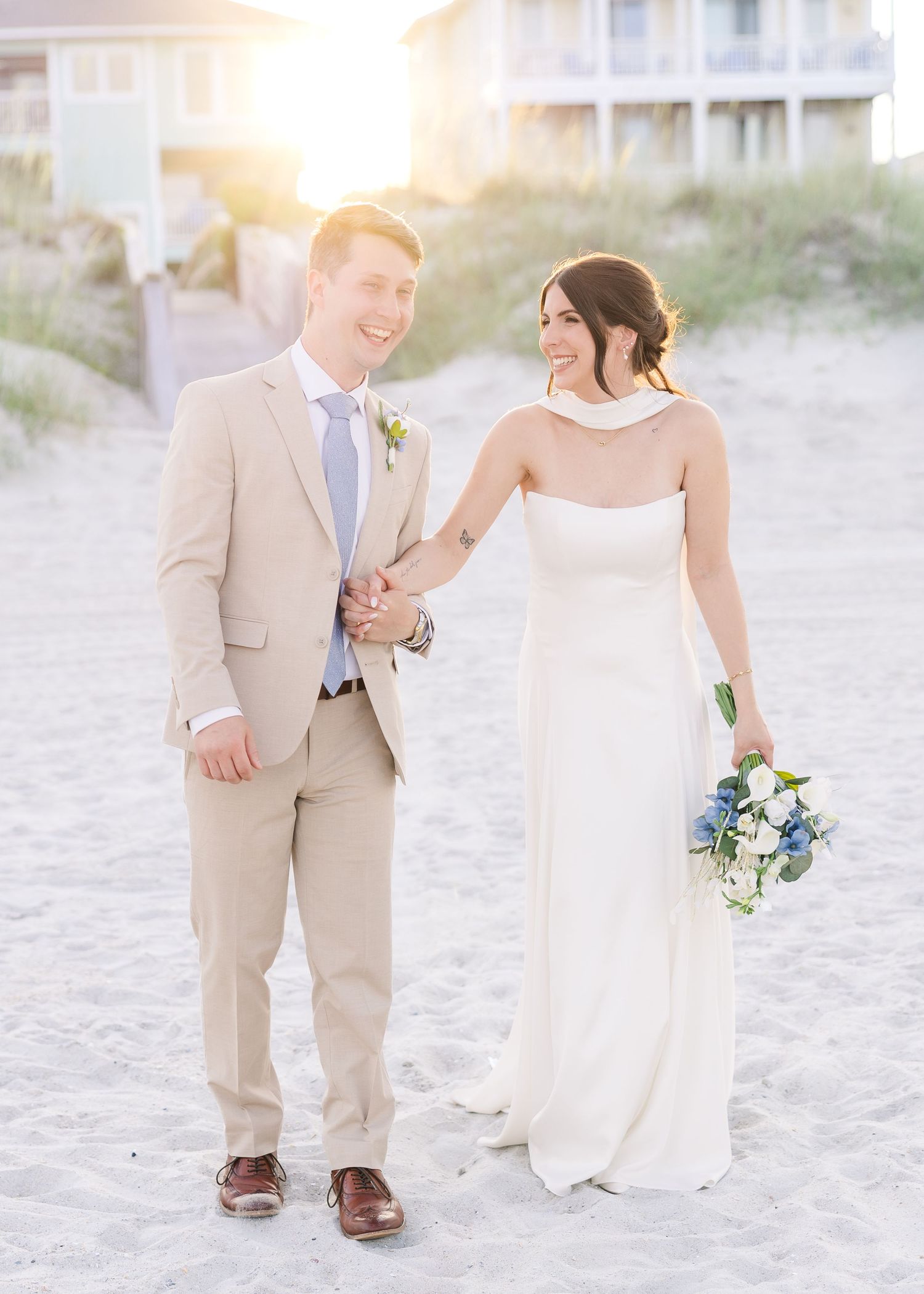 Bride and groom hold hands on beach at golden hour, groom in tan suit, bride in white gown in Carolina Beach near Wilmington, NC.