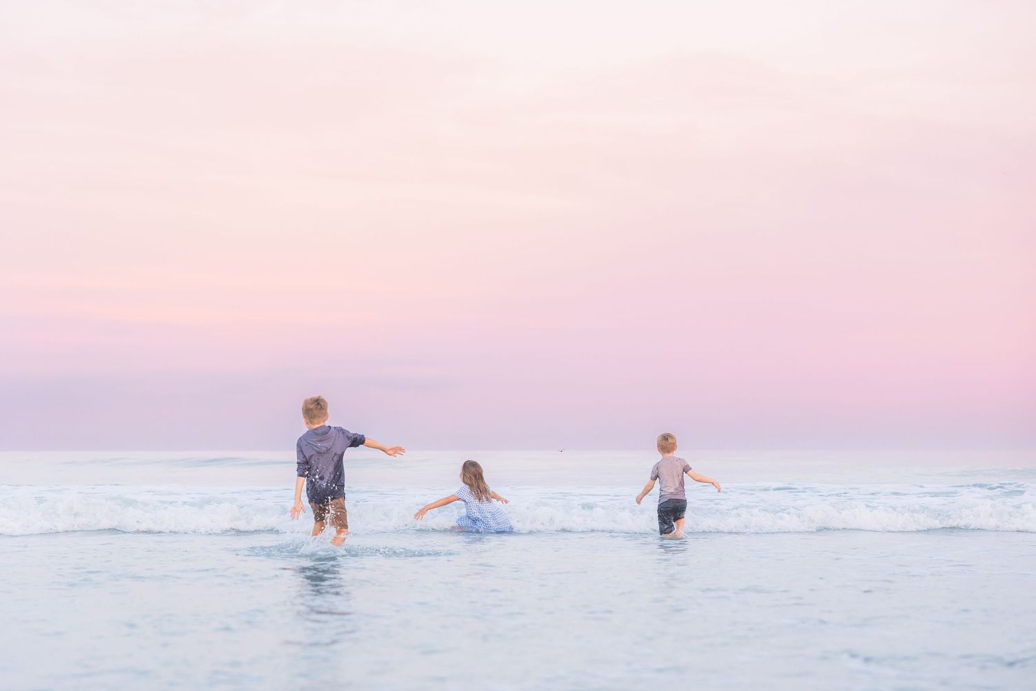 Children playing in gentle ocean waves during a pastel pink sunset.