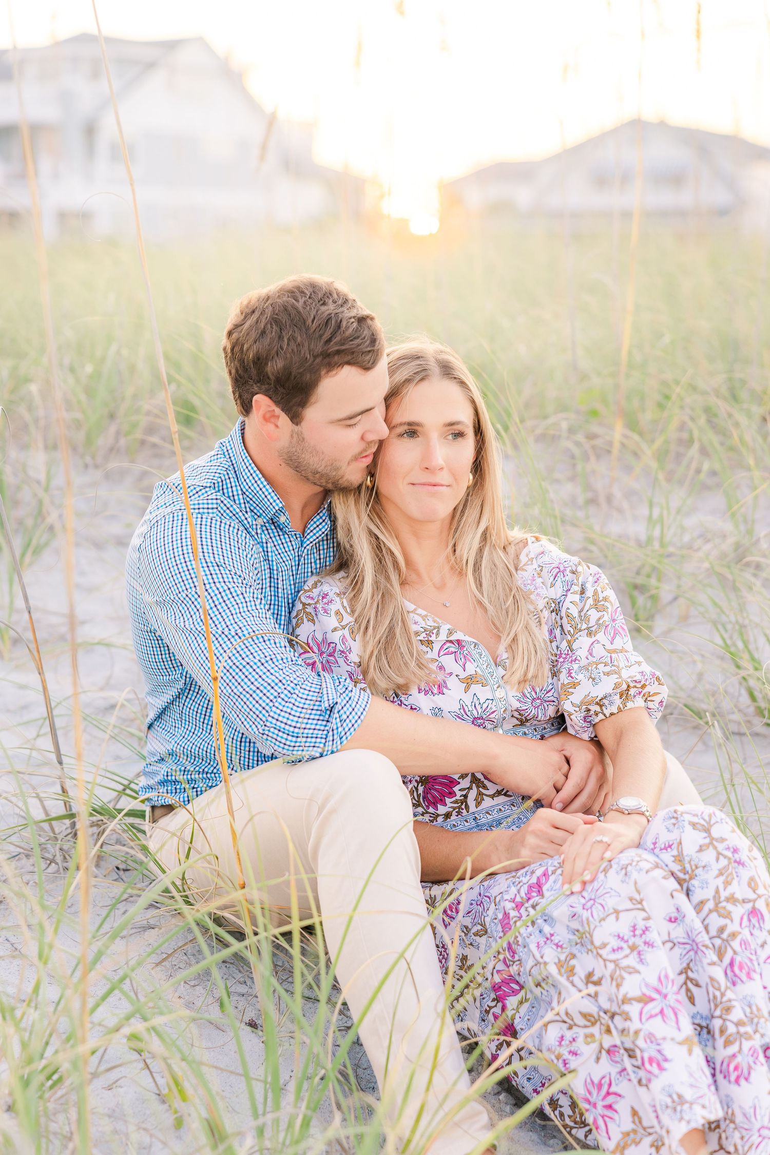 Intimate moment shared on sandy dunes with sunset and beach homes in distance.