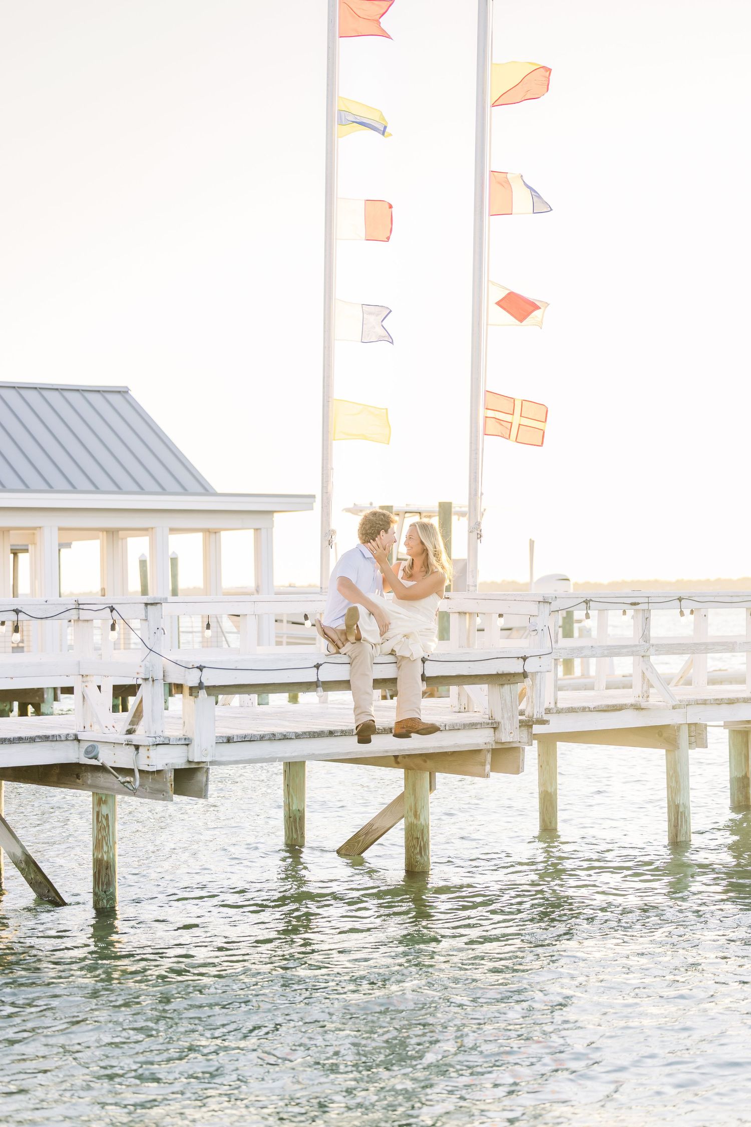 A couple shares a romantic moment on a pier with colorful flags and water in the background.