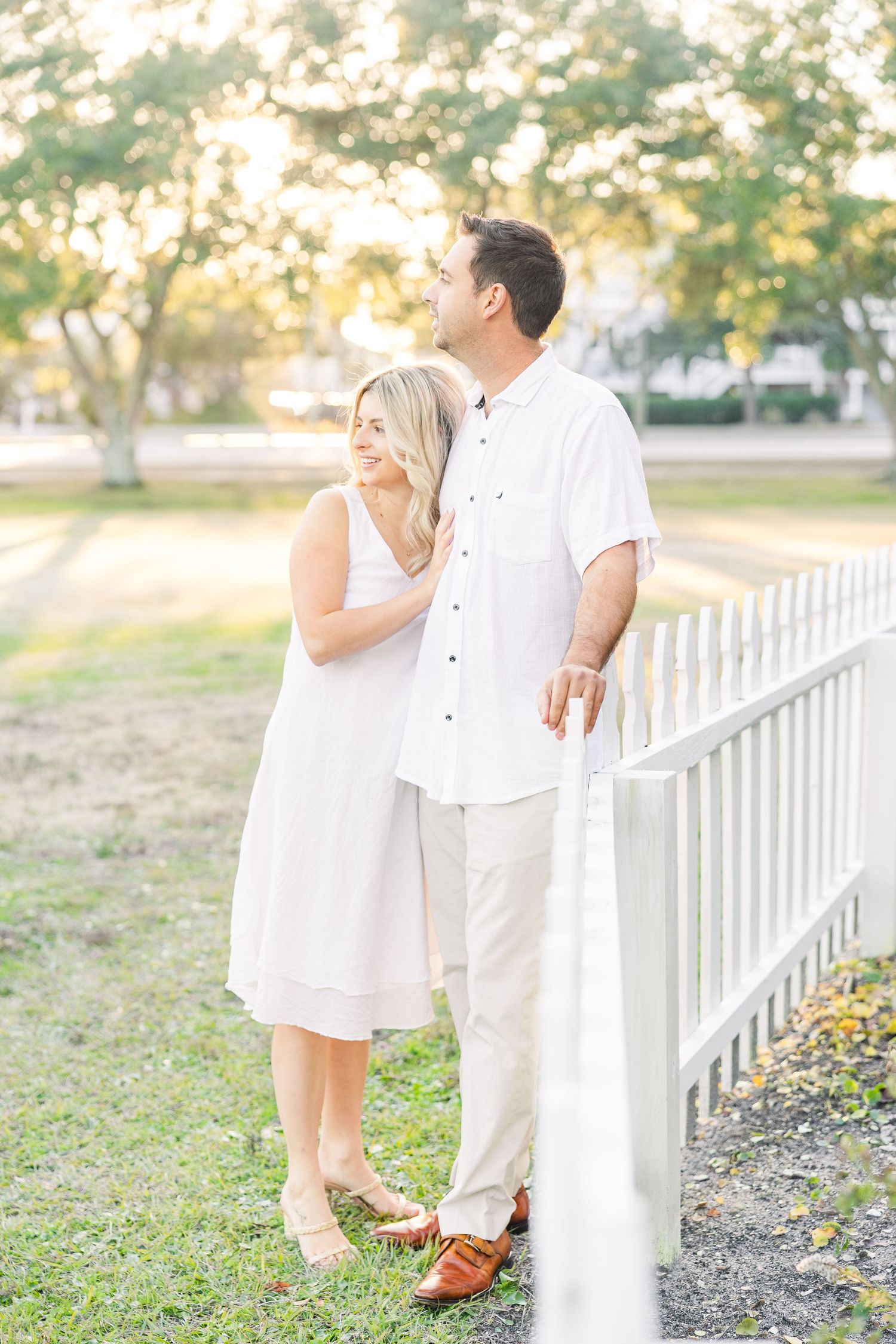 A couple in matching white outfits poses romantically by a white picket fence at sunset.