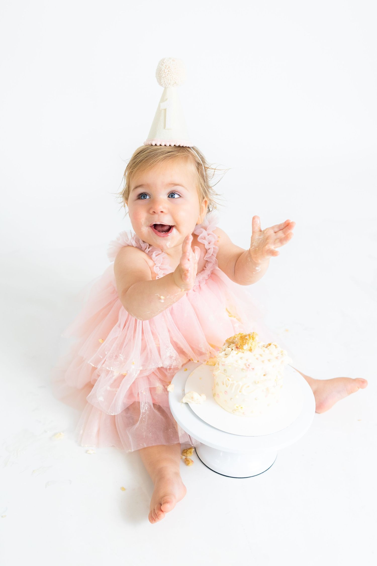 Excited baby in pink dress and party hat reaching up during first birthday cake smash photoshoot.