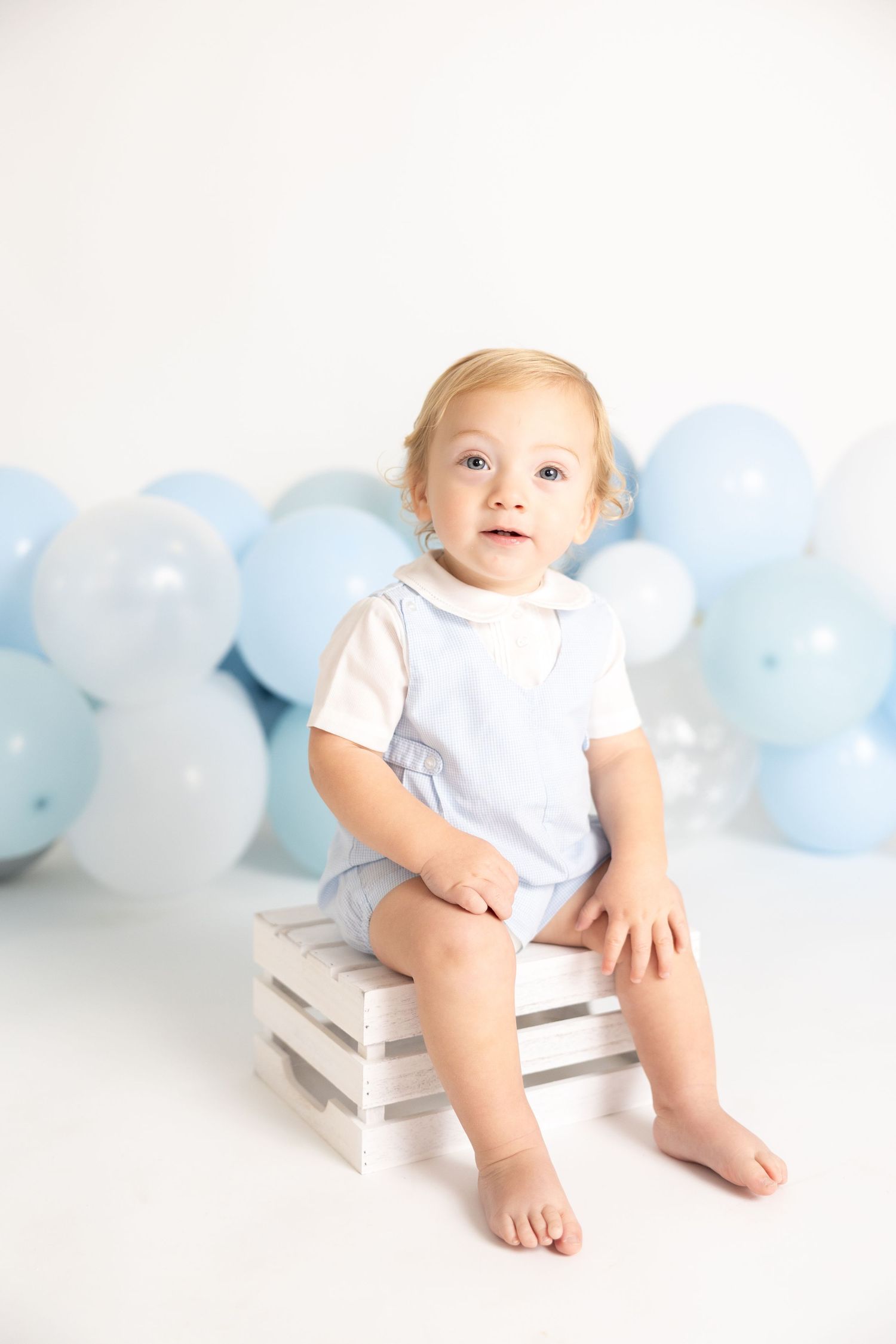 Baby in white outfit perched on white books with blue balloon backdrop.