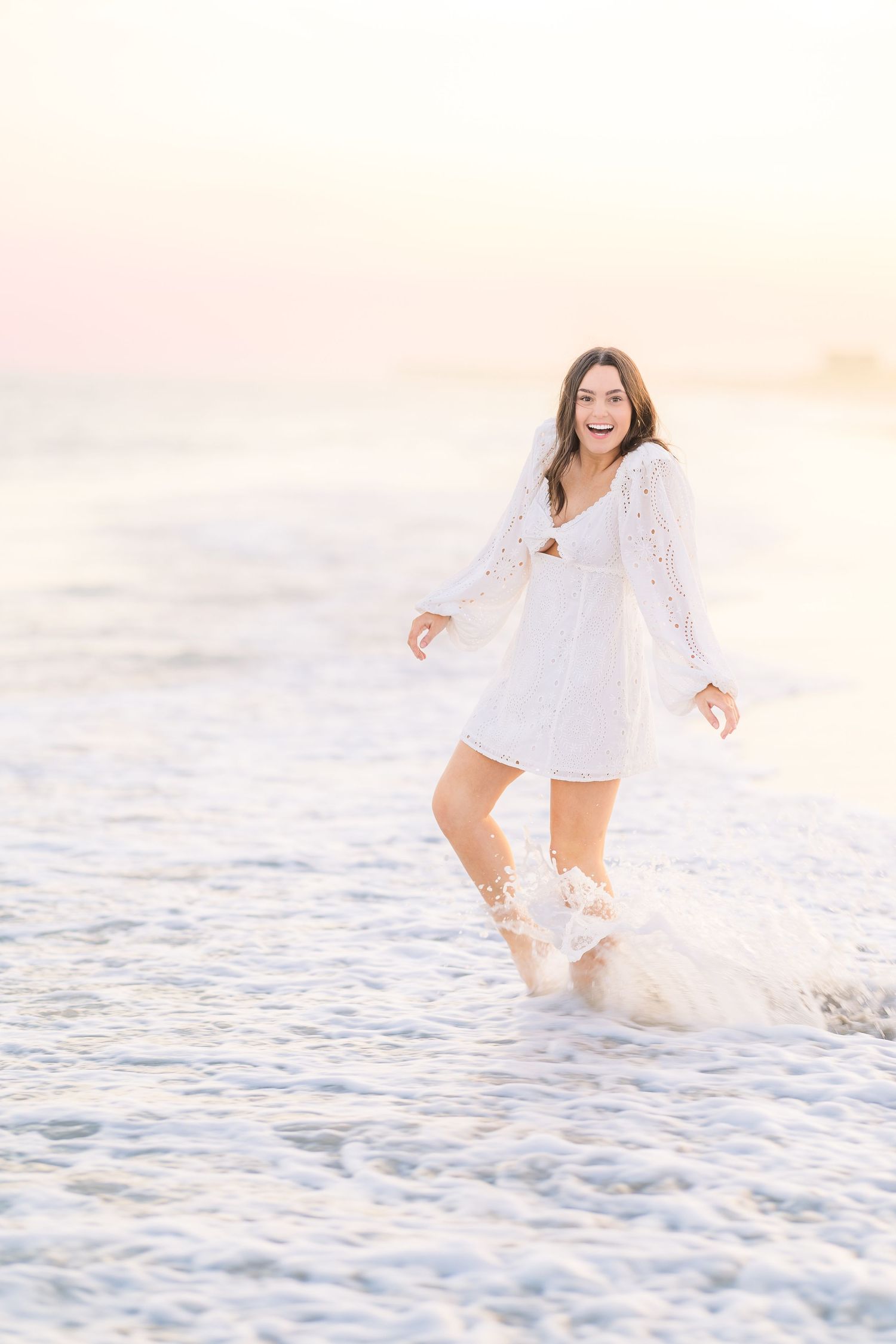 Joyful beach photoshoot featuring a white eyelet dress and splashing waves at sunset.