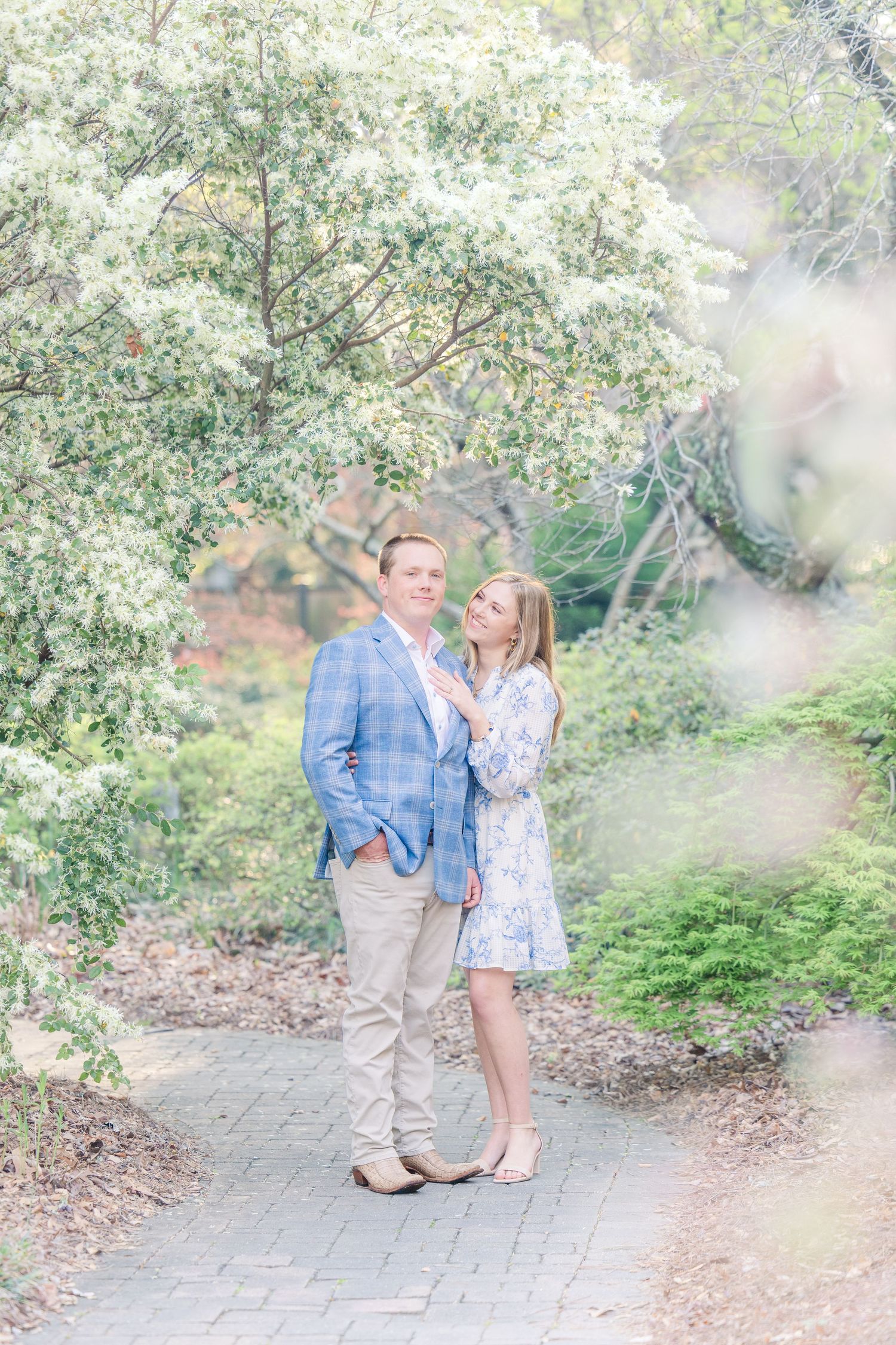 A couple stands together on a garden path surrounded by blooming white spring trees and soft natural lighting.
