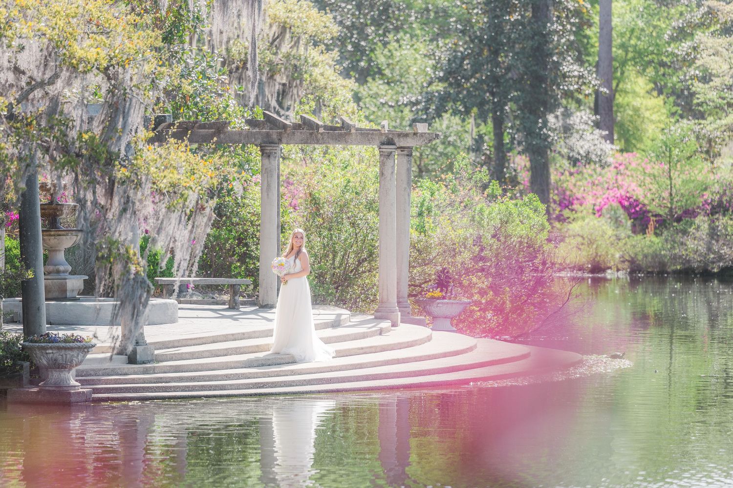 Bride in white gown stands on scenic garden gazebo steps surrounded by colorful blooms reflected in calm water Airlie Gardens in Wilmingto