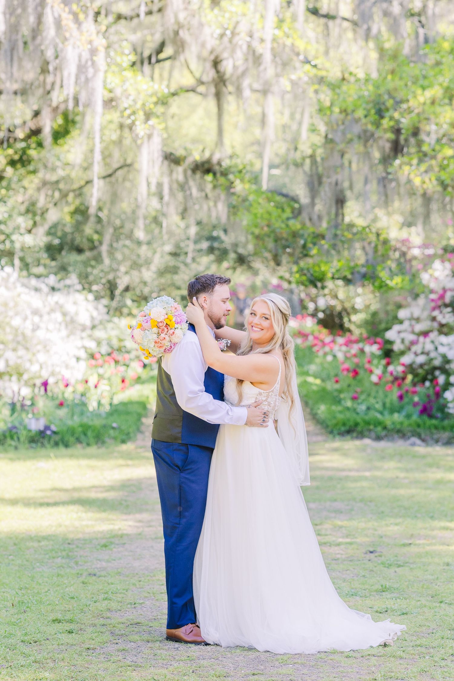 Bride and groom embrace in a lush garden with blooming flowers and Spanish moss-draped trees on their wedding day.