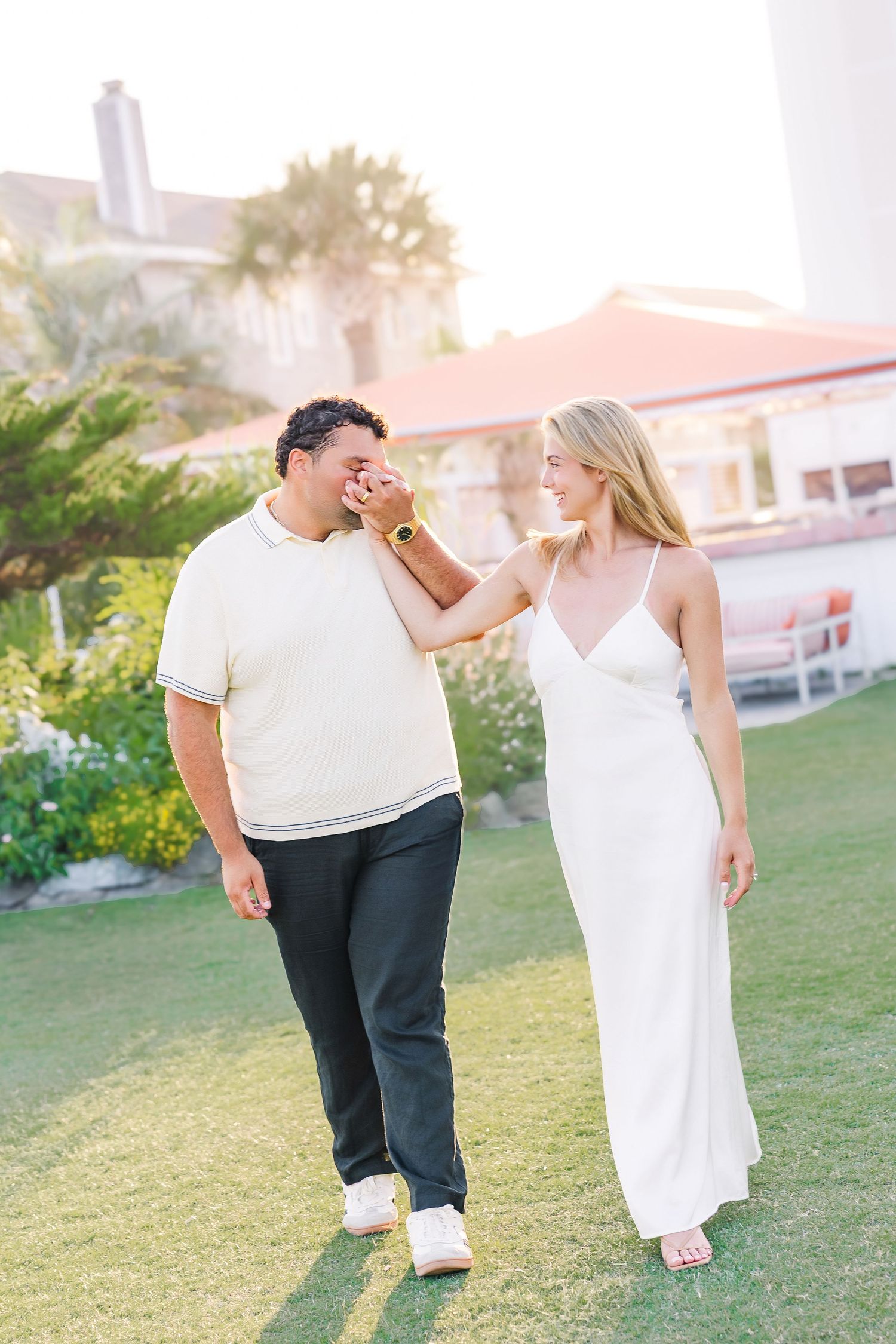 Happy couple walking together on resort lawn, woman in white dress touching mans face at sunset.