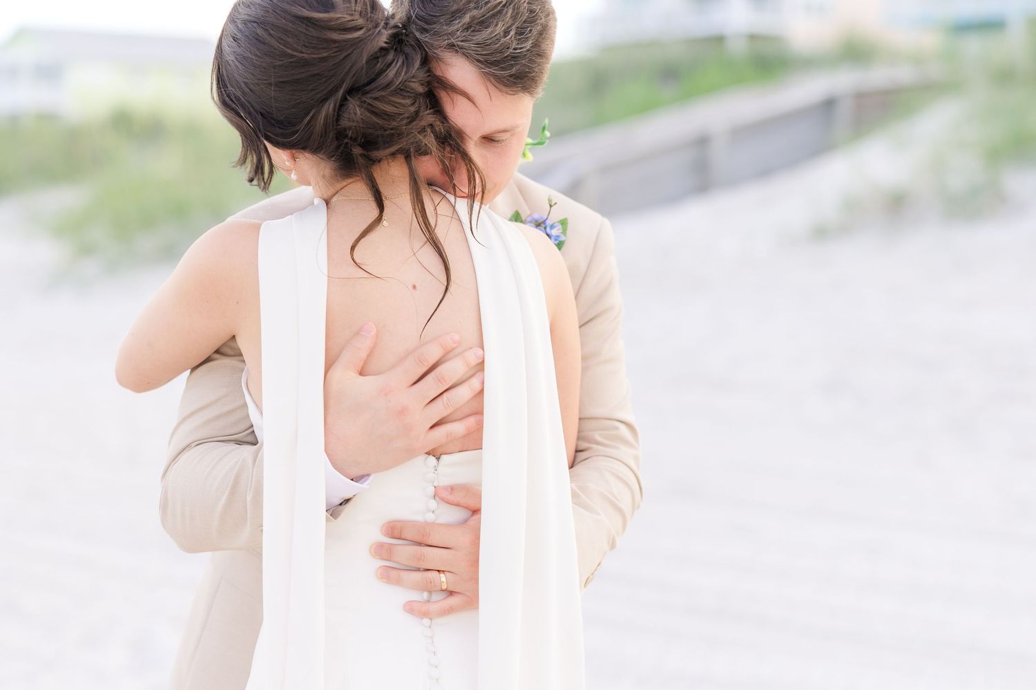 Groom tenderly embracing bride from behind on sandy beach during romantic wedding photo session in Carolina Beach near Wilmington, NC.
