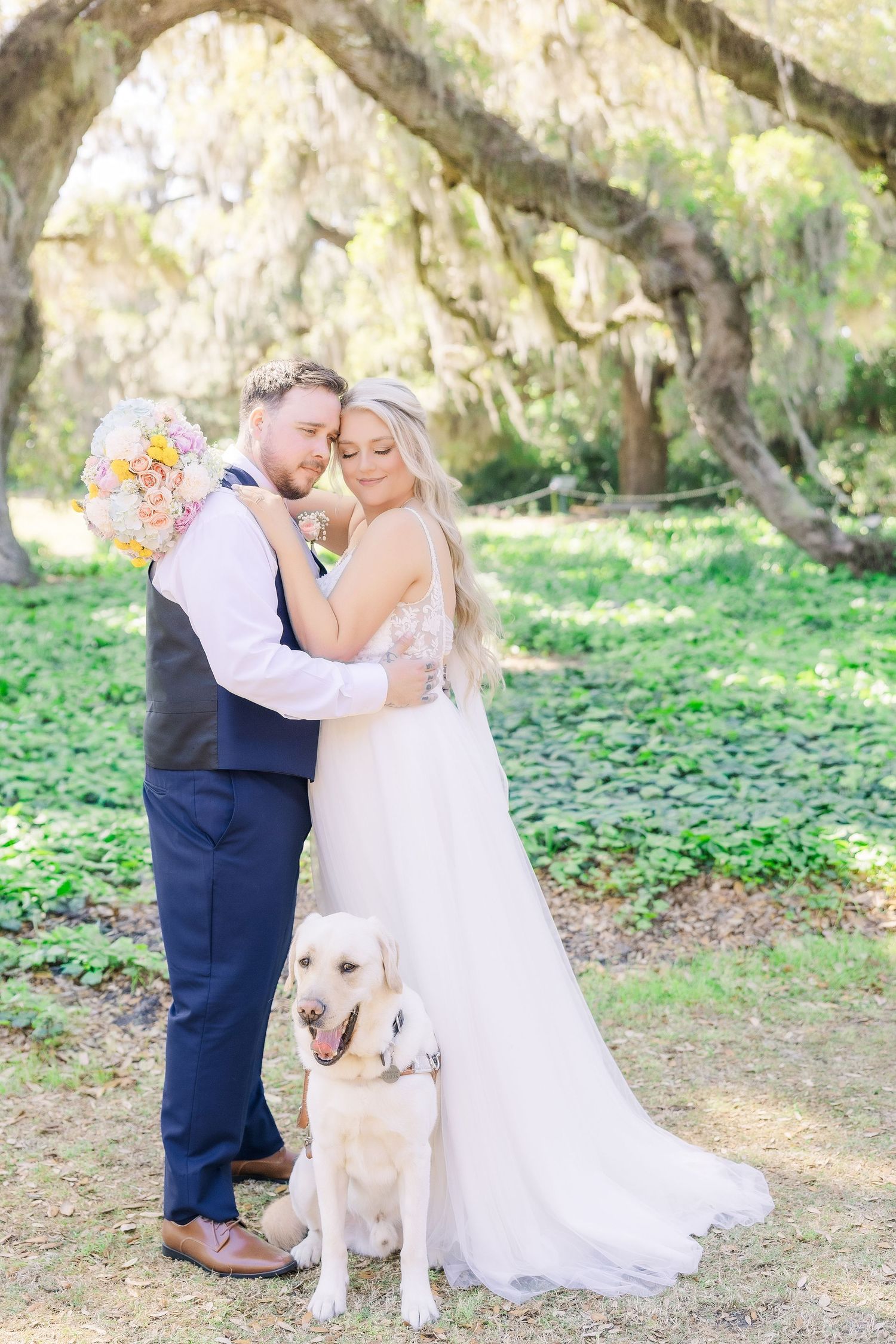 Bride and groom embrace under moss-draped oak trees with golden Labrador retriever at outdoor wedding in Airlie Gardens in Wilmington, NC