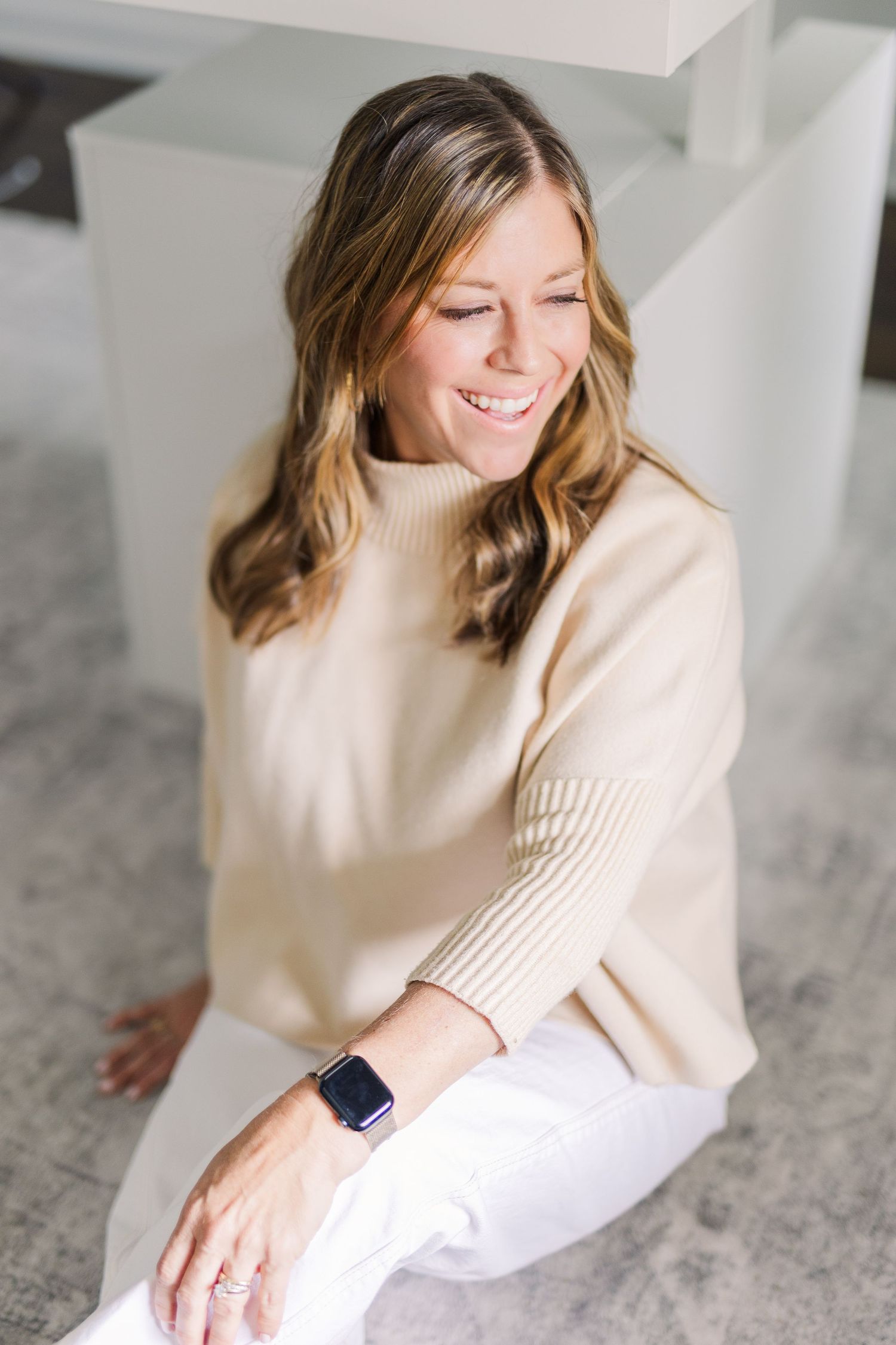 Lifestyle portrait of someone in cream turtleneck sweater with smart watch sitting against grey wall.