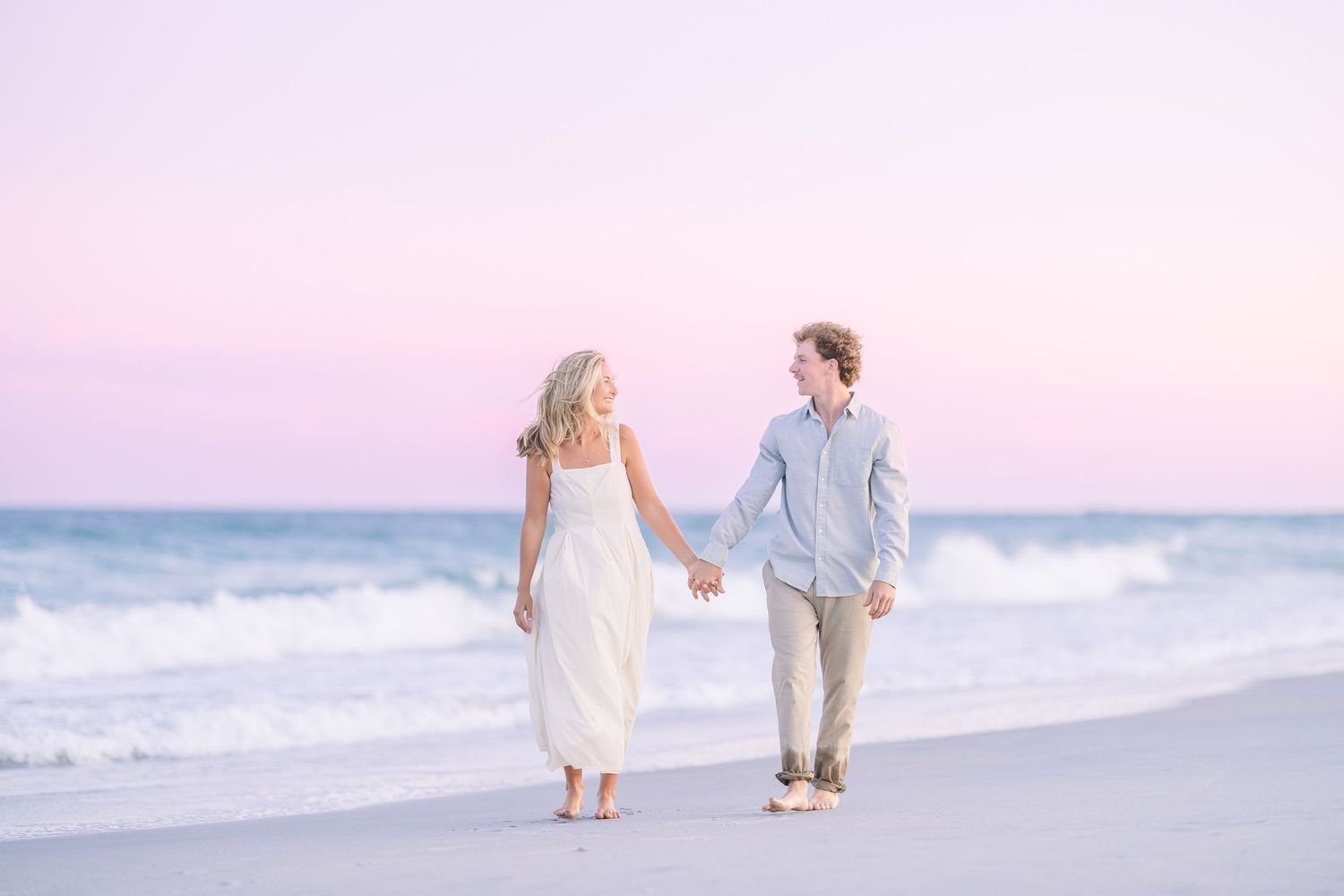 A couple takes a romantic stroll along the shoreline during golden hour with soft pink sky.