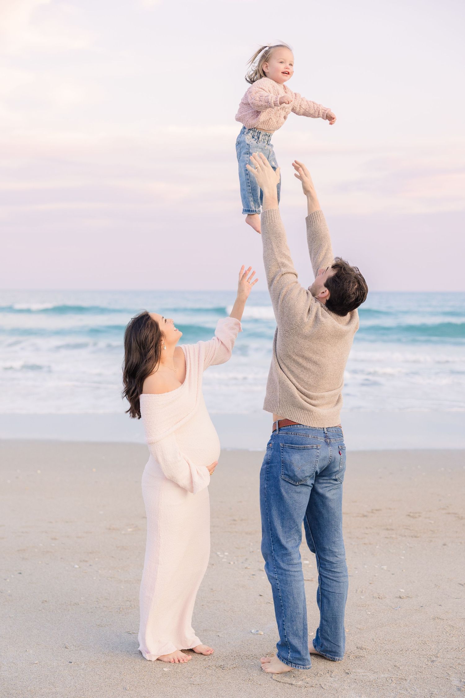 A family plays together on a peaceful beach at sunset, lifting a child in the air with joy.