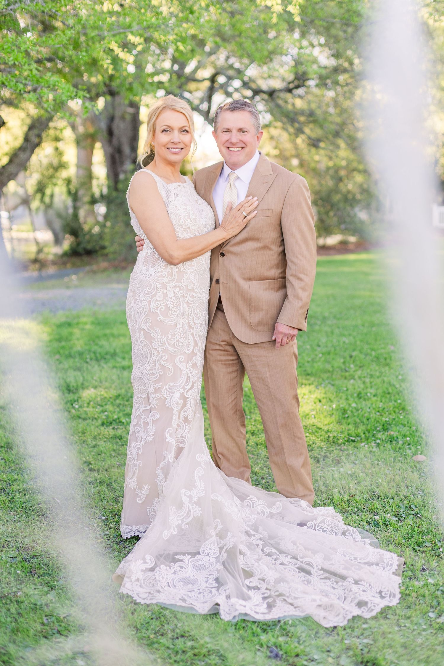 Couple poses on grass wearing tan suit and lace wedding gown with long train.