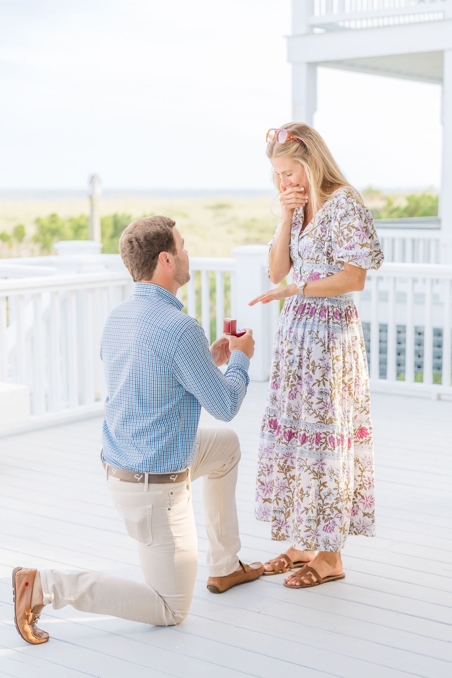 An emotional proposal moment unfolds on a white beachfront deck during daytime.
